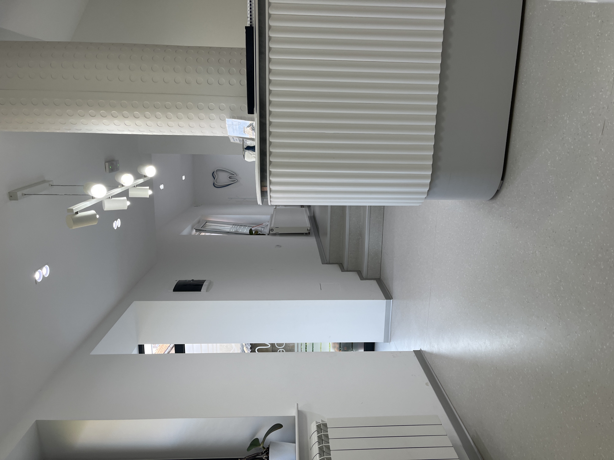 The image depicts the interior of a modern dental clinic located in Cacak, Serbia. The scene is a well-lit hallway with a reception desk on the right. The walls are painted white, and the floor is a light gray speckled material. To the left, a white radiator is visible below a window. Further down the hallway, stairs ascend to the right. A prominent feature is a white reception desk with a fluted front and a gray base. Above the desk, a cylindrical column with a textured, circular pattern rises towards the ceiling, where a contemporary multi-arm light fixture is suspended. A stylized blue tooth logo is mounted on the wall further down the hallway, confirming the establishment's purpose. No individuals are present in the frame.