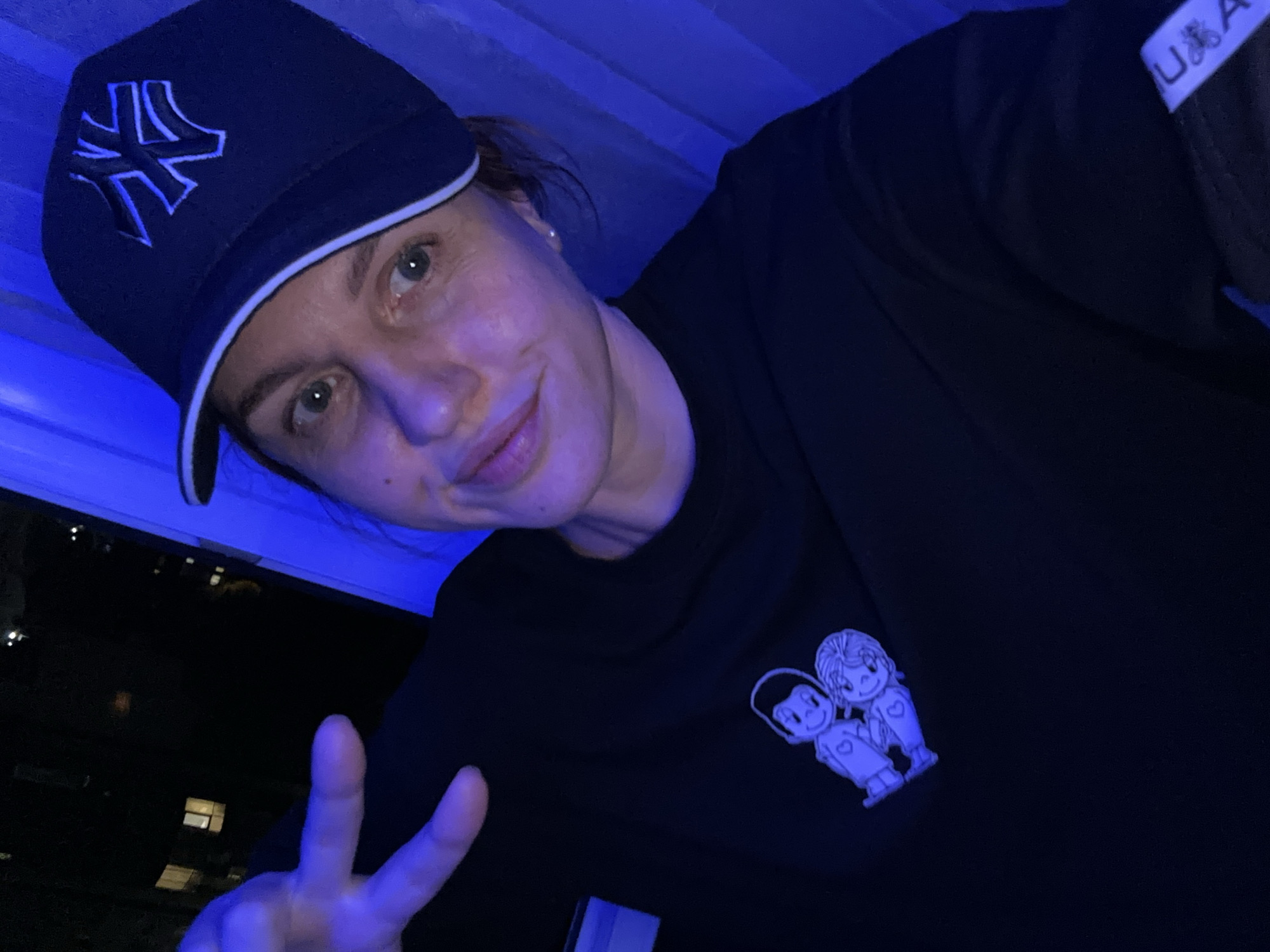A woman is taking a selfie in front of a window at night. She is wearing a black baseball cap with a white New York Yankees logo and a black t-shirt with a cartoon couple graphic. Her left hand is raised, making a peace sign. The background shows a dark city skyline with some illuminated windows, suggesting an urban environment. Blue lighting from an unseen source casts a strong glow on the scene, particularly on the right side of the woman's face and the window area. The woman's expression is neutral. There are no other people or animals visible in the frame. The image does not contain information to identify the location as Sochi, Russia.