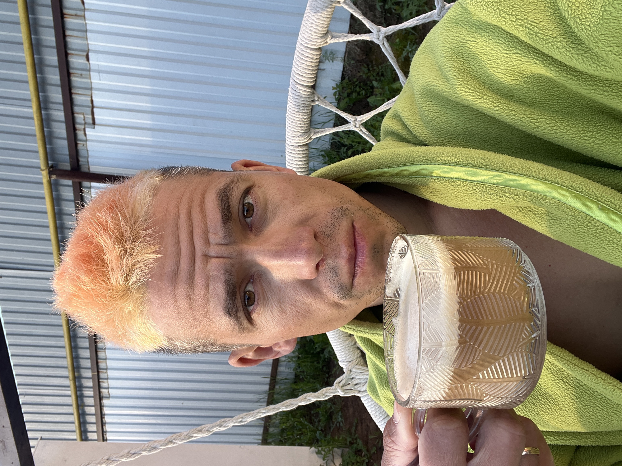A man with dyed orange hair sits in a white rope chair outdoors, wearing a lime green robe. He is holding a glass mug filled with a foamy beverage. The setting appears to be a backyard, with a corrugated metal fence visible in the background, possibly in the city of Chaikovsky, Russia.