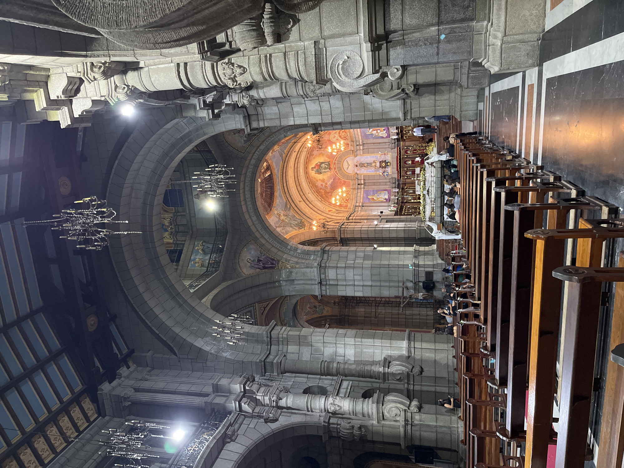 The image shows the interior of a large, ornate church, likely a cathedral, in Mérida, Venezuela. The architecture is grand, with high vaulted ceilings, stone arches, and intricately carved pillars. Several large, multi-tiered chandeliers hang from the ceiling, illuminating the space with a warm, golden light.
In the foreground, rows of polished wooden pews are arranged facing an altar. Some pews are occupied by individuals, while others are empty. The floor is a dark marble with white and orange stripes.
Further into the church, the altar area is visible. It is elaborately decorated with religious iconography, including statues and paintings. Several people are gathered around the altar, some dressed in what appear to be clerical garments, suggesting a service or ceremony is taking place. The overall atmosphere is one of solemnity and reverence. The specific location is identified as the Basilica Menor de la Inmaculada Concepción, Mérida, Venezuela.