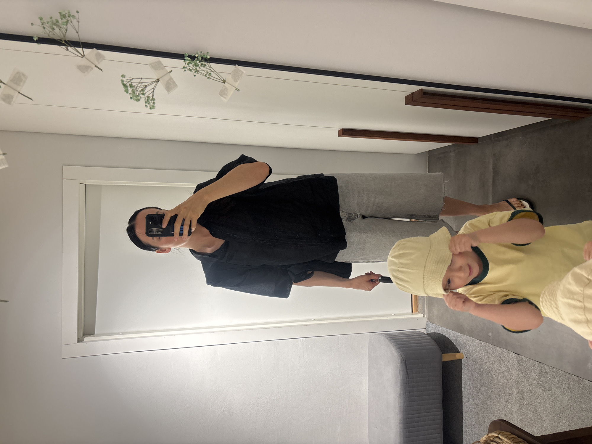 In a brightly lit interior space, a mirror reflects a scene featuring two individuals. A woman in a black top and grey shorts is taking a photo of herself and a young child. The child wears a light yellow shirt with dark green trim and a matching bucket hat. The environment appears to be a modern room with white walls, grey flooring, and a grey bench. Flowers are taped to the mirror frame. The location is not discernible from the image.