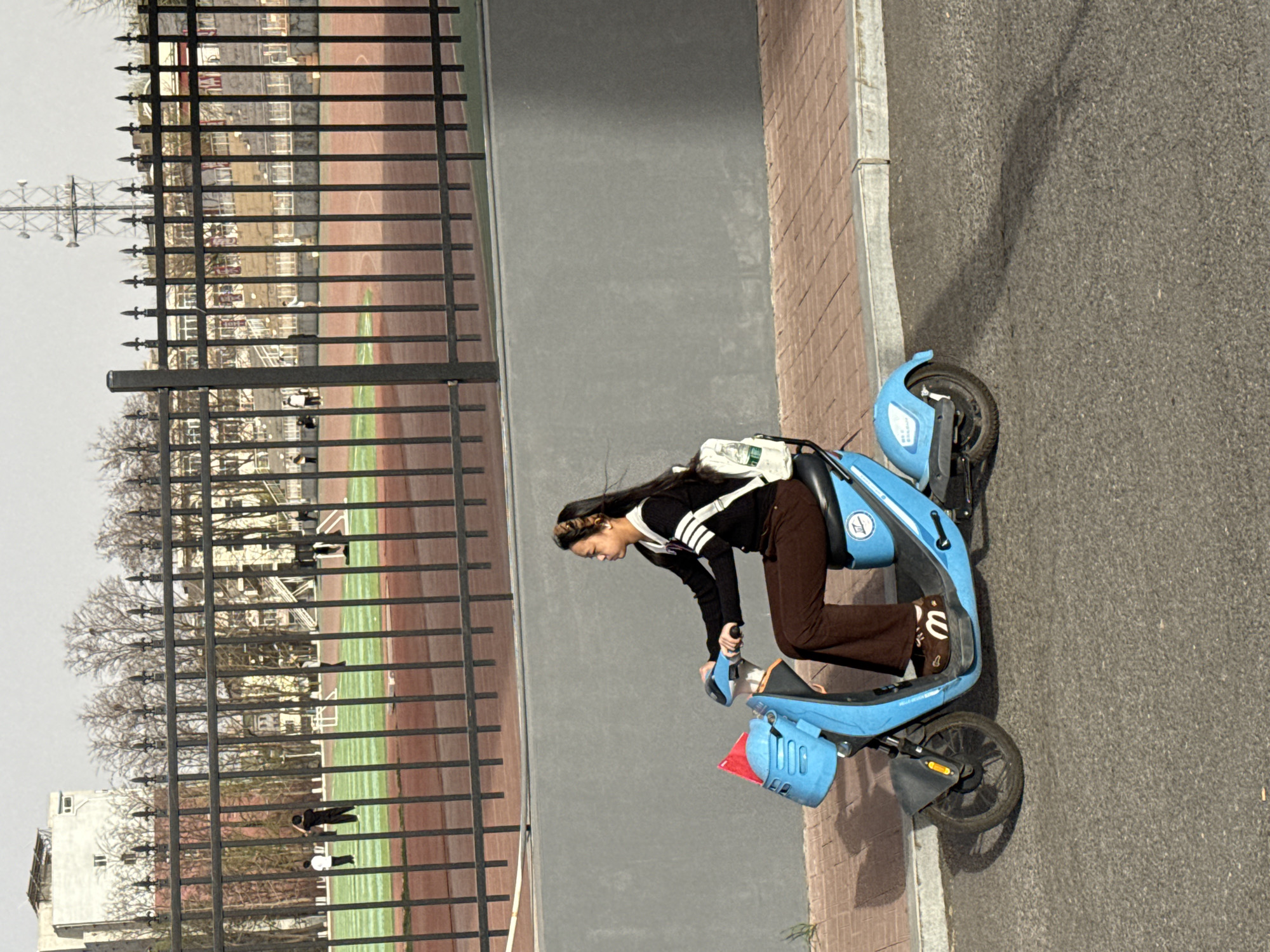 A young woman rides a light blue electric scooter on a sidewalk. The scooter is positioned on the right side of the frame, angled slightly away from the camera. The woman is wearing a dark, long-sleeved top with white stripes, brown pants, and brown slip-on shoes with white lettering. She has long, dark hair. A white backpack is slung over her shoulder.
Behind the sidewalk and the woman is a low concrete wall, and behind that is a metal fence, beyond which is a sports field. Some people are visible on the field, and there are trees and buildings in the background. The overall setting suggests a city environment, specifically Zhongtun, China, as referenced in the prompt. The weather appears to be overcast.