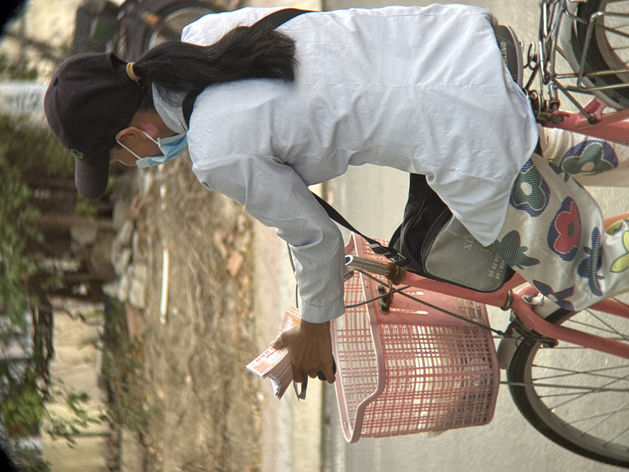 A person wearing a black baseball cap, a light blue face mask, and a light blue striped shirt is riding a pink bicycle. The person is holding a stack of pink lottery tickets in their right hand, which is extended over a pink wicker basket attached to the front of the bicycle. The person is wearing floral patterned pants. The bicycle has a black bag attached to the handlebars and a rack on the back. The background shows a dirt road and vegetation. The image appears to have been taken on a sunny day. The context of a person selling lottery tickets from a bicycle is common in urban areas of Vietnam, including Nha Trang.