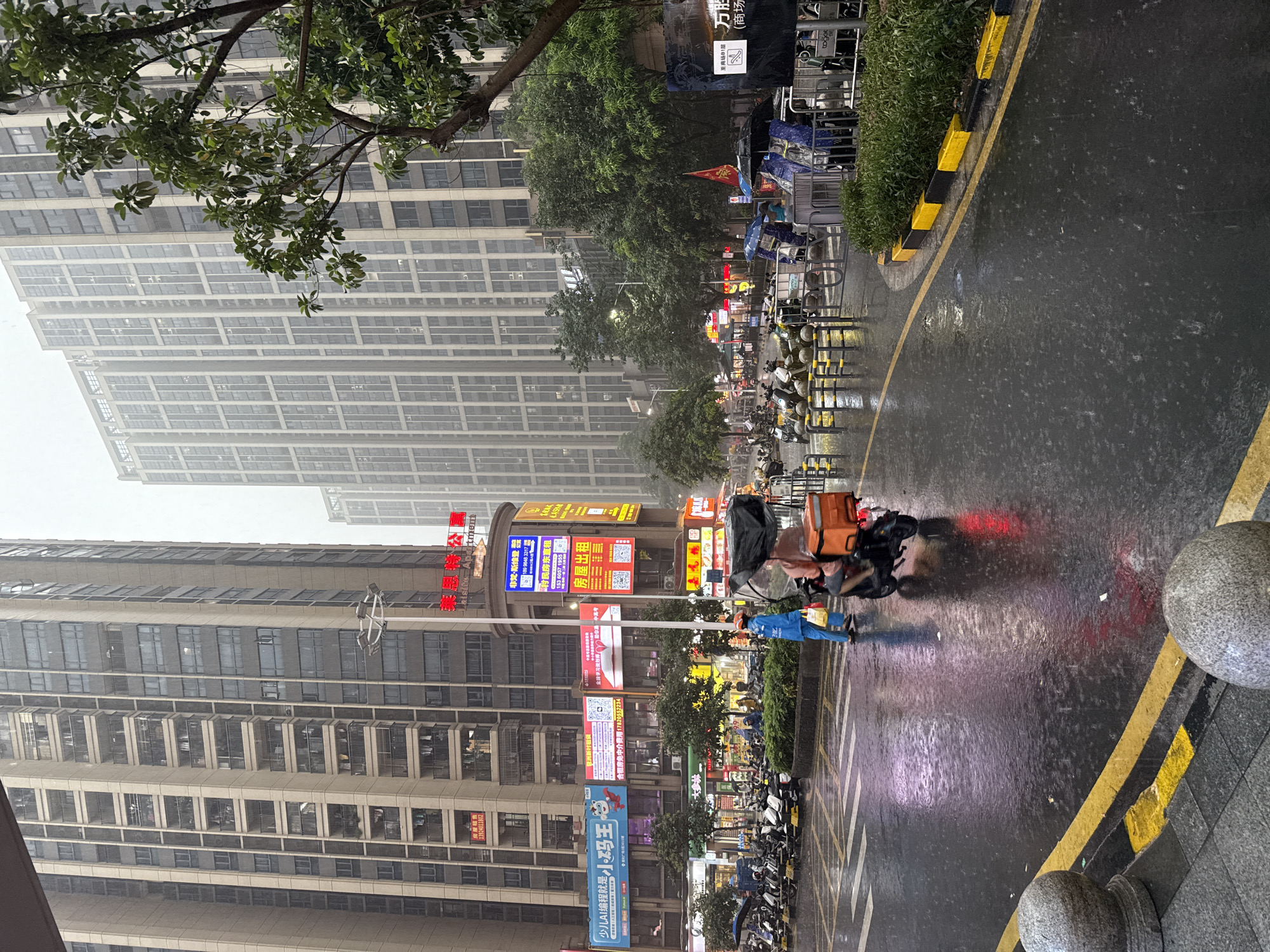 The image depicts a street scene in Haizhu District, China, during a downpour. Two tall, modern apartment buildings dominate the background, their facades a grid of windows and balconies. In the midground, a street is wet and reflective from the rain, with numerous parked motorcycles. A delivery person on a scooter, wearing a blue uniform and carrying an orange insulated bag, is in the process of riding away, the umbrella over their head providing minimal protection. Another person, also under an umbrella, is walking nearby. The street is lined with shops and illuminated by neon signs, some advertising apartments for rent and other services. Trees with lush green foliage are visible, adding a touch of nature to the urban environment. The overall atmosphere is one of a typical city day, made more dramatic by the heavy rain.