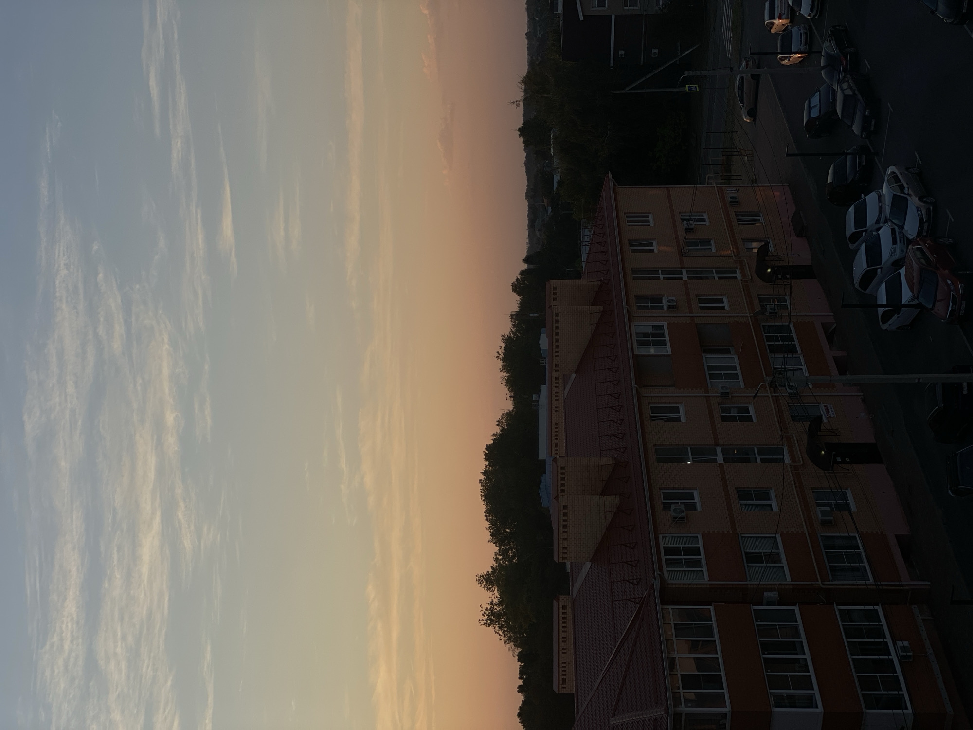 The image captures a residential area in Krasnodarskaya Ptitsefabrika, Russia, during what appears to be dusk. The sky is a gradient of pale blue and soft orange, with wispy clouds stretching across the horizon. In the foreground, a multi-story brick apartment building with a red tiled roof is visible, featuring numerous windows and balconies. Adjacent to the building, a parking lot is filled with several cars of various models and colors. Further in the background, a dense line of trees separates the residential area from other structures, likely more buildings and some distant greenery. The overall scene is quiet and still, bathed in the fading light of the day.
