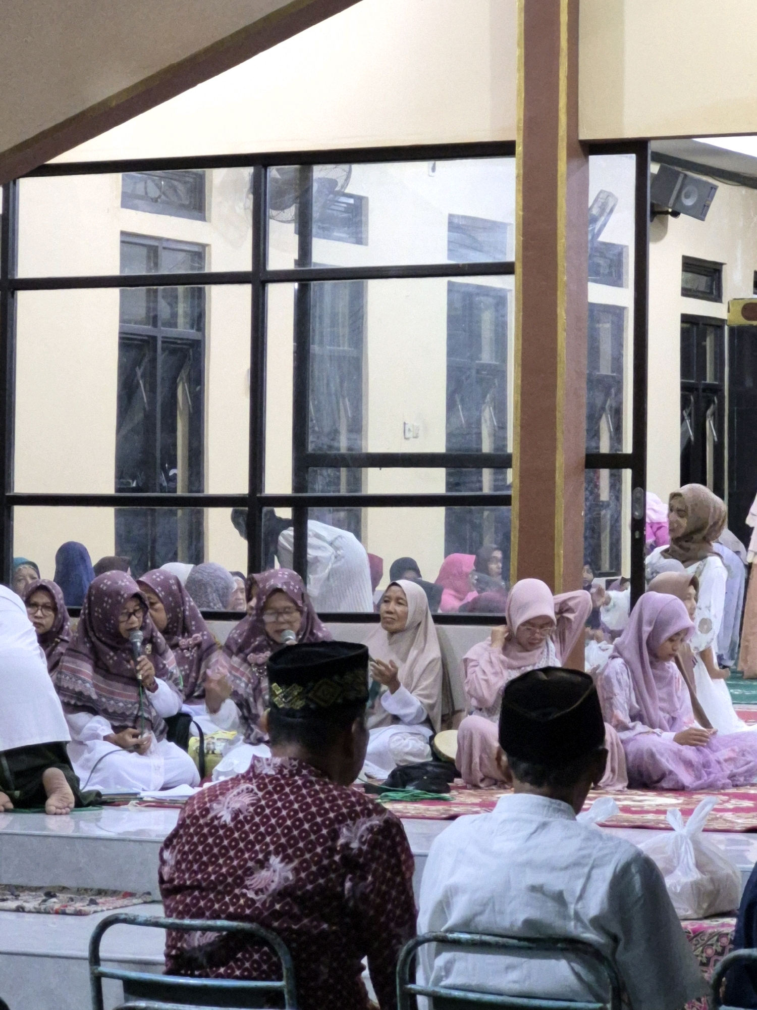A group of people, mostly women wearing hijabs and headscarves in various shades of purple, pink, and beige, are seated on the floor in a room that appears to be a community hall or place of worship. The women are facing a microphone, suggesting they are engaged in a discussion or presentation. One woman in the foreground on the left is speaking into a microphone. To the right, two men are seated, one in a patterned maroon shirt and a black peci, and another in a white long-sleeved shirt. They are facing away from the camera, looking towards the group of women. The environment is characterized by beige walls, large grid windows with reflections of the building's interior and exterior, and wooden beams with gold accents. In the background, through the windows, a building with multiple stories and windows is visible. A fan is mounted on one of the exterior walls. The lighting appears to be artificial and somewhat dim, casting a warm glow on the scene. The floor is covered with a patterned rug. The presence of plastic bags on the floor suggests a gathering or event. The overall scene depicts a religious or community gathering in Dukuhrejo, Indonesia, likely a mosque or community center, given the attire and the focus on religious discourse.