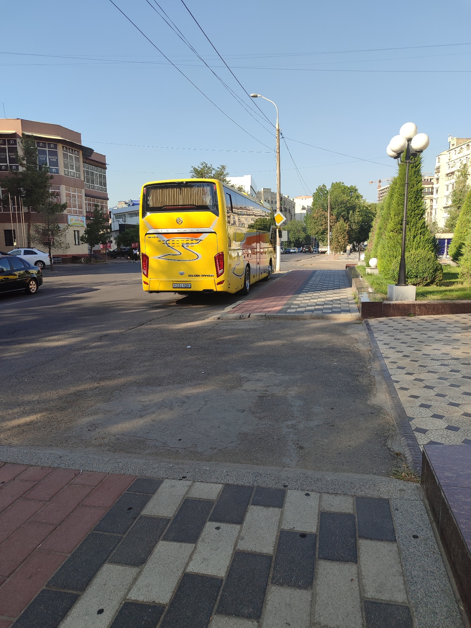 A bright yellow bus with the inscription "GOLDEN DRAGON" on the back is parked on the right side of a city street in Tashkent, Uzbekistan. The bus is viewed from the rear, and its license plate, "HG314 ADA," is visible. To the left of the bus, a black sedan is parked on the side of the road. Across the street, a multi-story building with a brown facade and numerous windows stands under a clear blue sky. A single street lamp is visible on the left side of the frame. The foreground features a sidewalk paved with a pattern of grey, black, and reddish-brown rectangular tiles. A decorative lamppost with three white globe-shaped lights stands on the right side of the image, adjacent to a row of neatly trimmed green trees. The scene appears to be an urban street on a clear day.