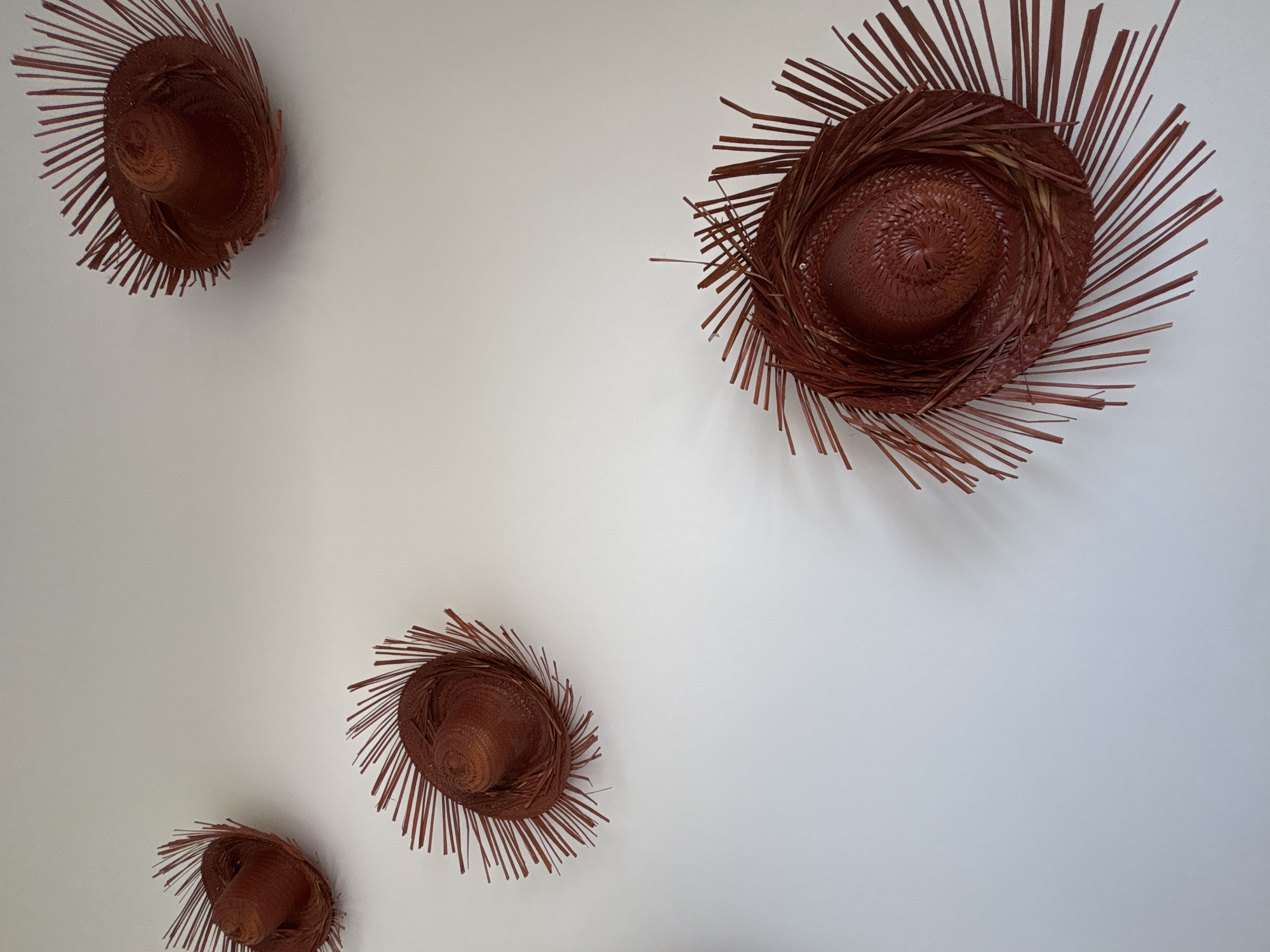 Four woven hats with radiating straw decorations are positioned against a plain white wall in Baan Chaweng Noi, Thailand. The hats vary in size and placement, with the largest one located in the upper right corner and the smallest in the lower left.