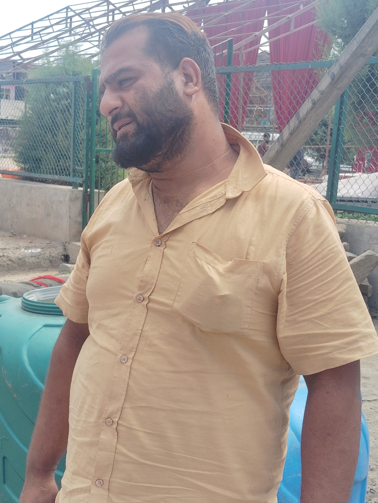 A medium shot photograph shows a man from the chest up, facing to the left and looking down. He has a full beard and short, dark hair. He is wearing a light tan, short-sleeved, button-down shirt with a collar. The shirt has a single chest pocket on the left side. The man stands in front of a green chain-link fence and a large teal plastic tank. Behind the fence, a red fabric canopy is visible, supported by a wooden beam. In the background, there are structures and trees, indicating an outdoor setting in Srinagar, India.
