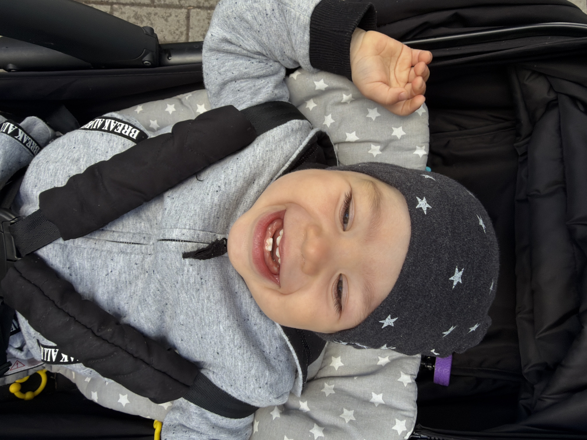 A joyful baby lies in a black stroller, looking upwards towards the camera with a wide, open-mouthed smile, revealing several small teeth. The baby's eyes are bright and engaged, conveying happiness. They are dressed in a grey hoodie with black cuffs, which has speckles of darker grey. A dark grey beanie adorned with white stars covers their head. One arm is raised above their head, and their hand is open.
The stroller's interior is lined with a soft, grey blanket or padding featuring a white star pattern. Black, padded straps secure the baby, suggesting a safe and comfortable ride. The stroller itself is black with visible handles and other components. Small, colorful objects, possibly toys or clips, like a purple one and a yellow one, are visible within the stroller. The background, partially visible, suggests an outdoor setting with concrete or pavement, consistent with Chaikovsky, Russia.
Visible text on a black strap across the baby's chest reads "BREAK AWAY". The overall impression is one of a happy infant enjoying an outing in possibly cooler weather, given the warm attire.