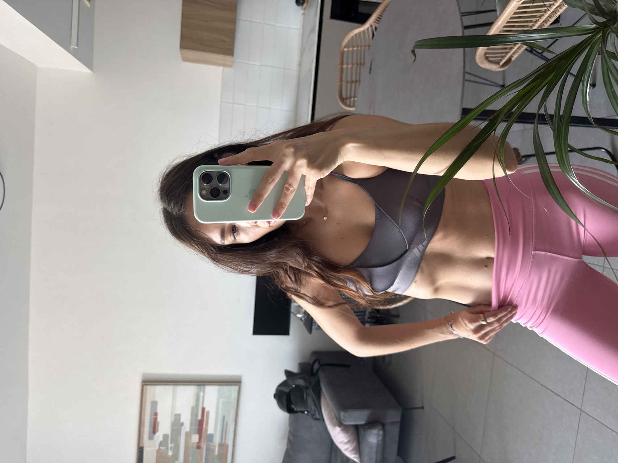 A woman is taking a selfie in what appears to be an apartment. She is wearing a gray sports bra and pink leggings. The apartment is modern with a couch, a picture on the wall, and a plant. There is no information to confirm this is Tel-Aviv, Israel.