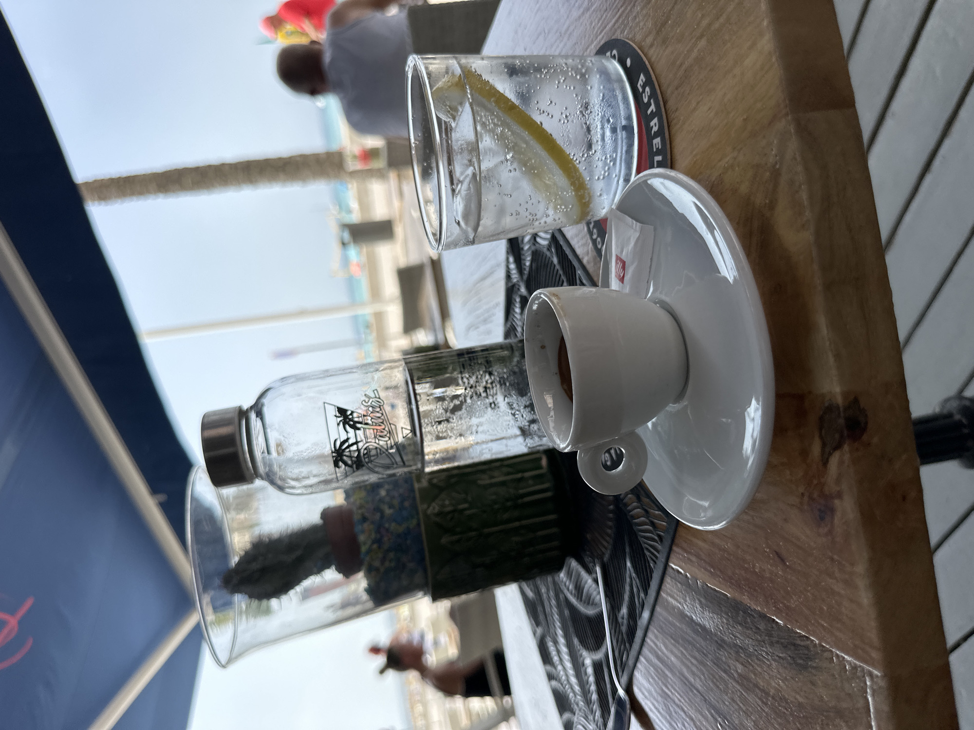 An outdoor cafe setting in Can Pastilla, Spain, features a wooden table with a coffee cup and saucer, a glass of sparkling water with lemon and ice, a decorative glass vase containing a cactus and colorful pebbles, and a clear glass water bottle with a metal lid. The table is adorned with a black placemat. In the background, blurred figures of people are visible, suggesting a public space near a beach. The sky is a bright blue, indicating daytime. The overall atmosphere appears relaxed and casual.
