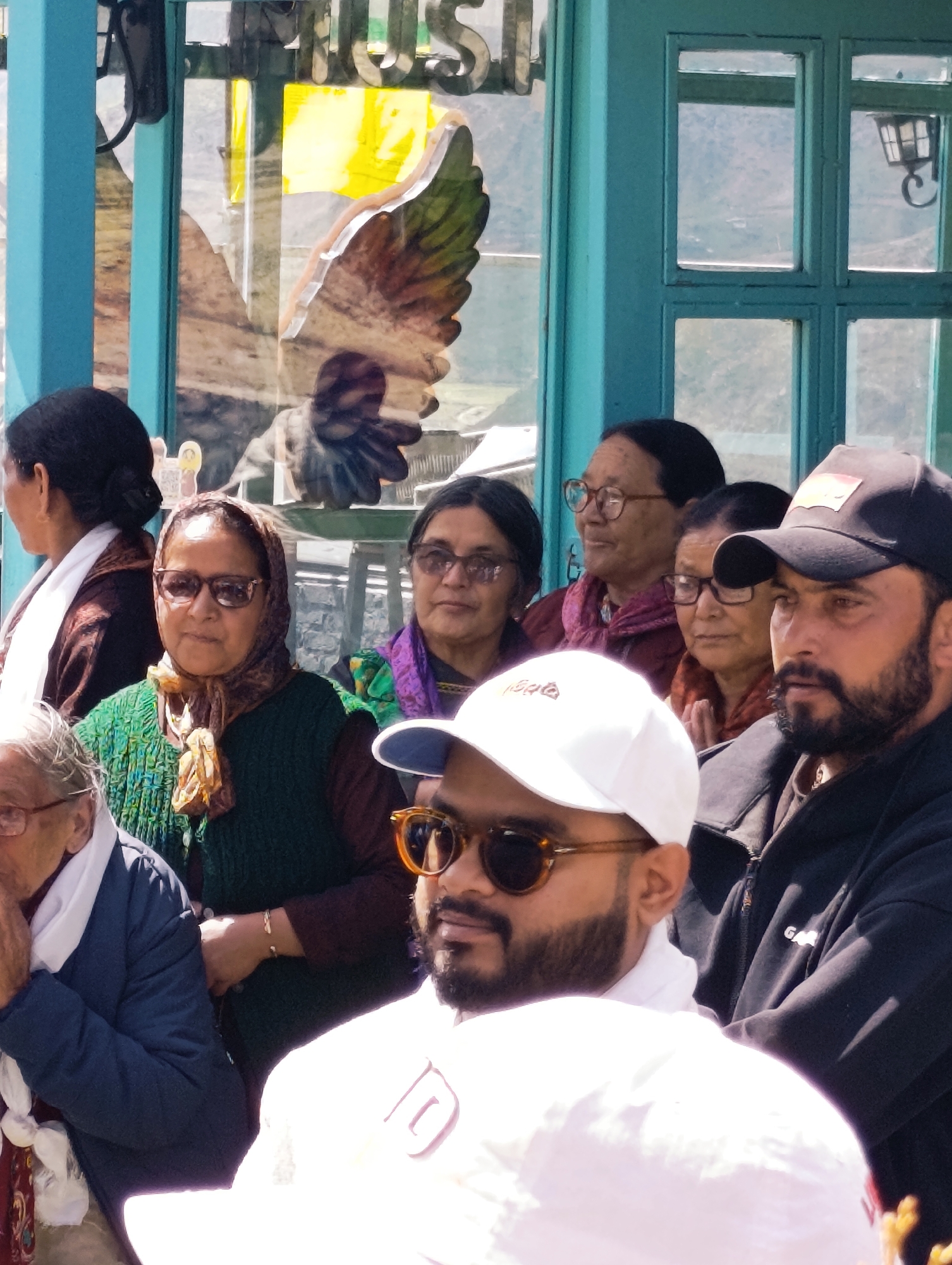The image depicts a group of people gathered outdoors in what appears to be a marketplace or scenic spot in Yamling, India. The background features a turquoise-colored structure with glass windows, suggesting a shop or cafe. Reflections in the glass show distant mountains and what might be a building. A large decorative element resembling a feathered wing is visible on the left side of the structure, possibly an artistic installation.
In the foreground, several individuals are visible. On the far left, a woman with dark hair is partially obscured. To her right, a woman wearing glasses and a headscarf is looking towards the viewer. Next to her, another woman with glasses and a colorful scarf is also looking forward. A man in a black baseball cap and sunglasses, with a beard, is prominently featured on the right side of the image, looking to his left. In front of him, a person wearing a white cap and sunglasses is also visible. The clothing of the individuals suggests a moderate climate. The overall scene conveys a casual gathering of people enjoying the outdoors in a scenic location.