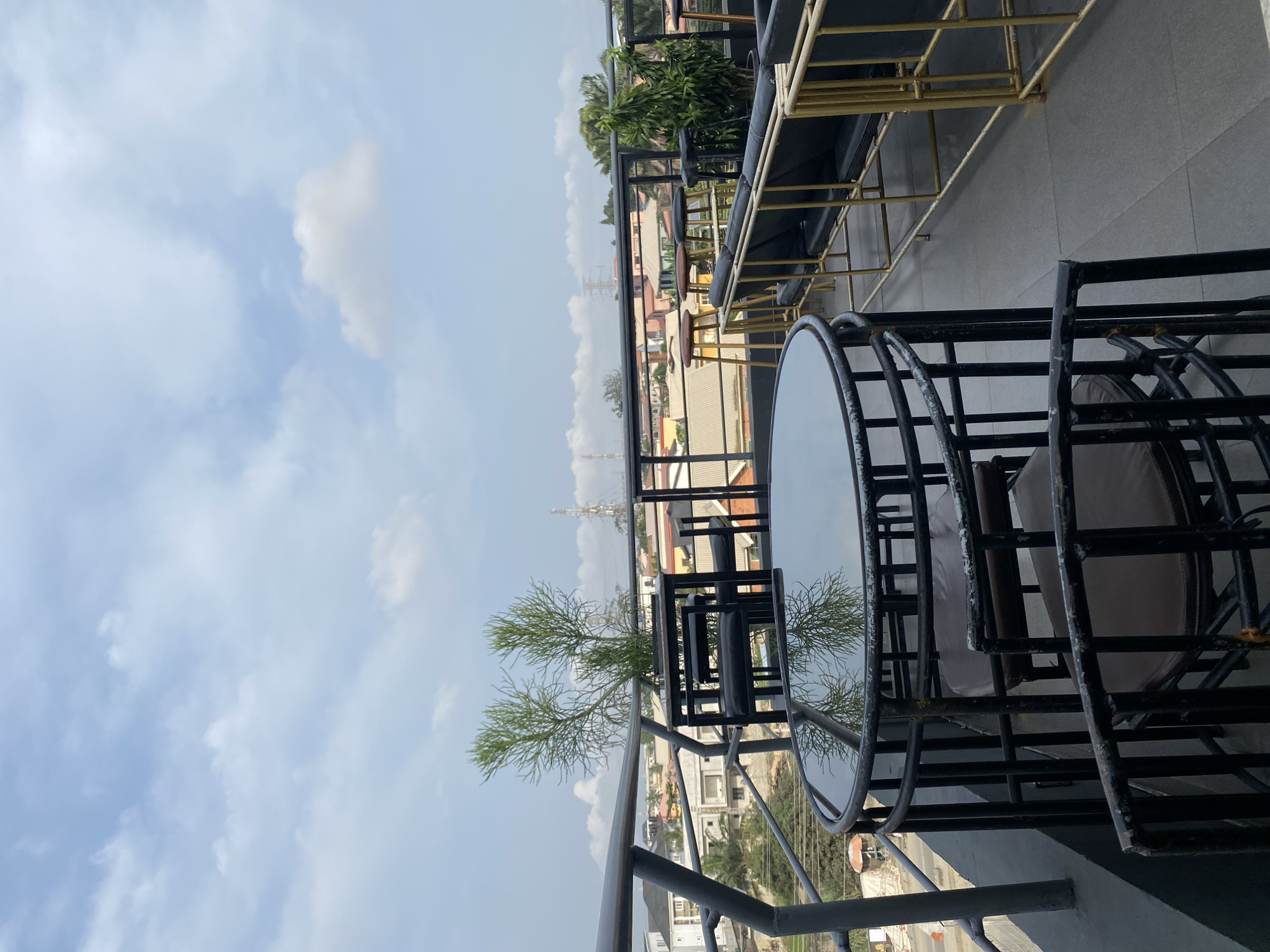 The image shows an outdoor rooftop seating area with a view of a city skyline under a cloudy sky. In the foreground, there is a round black metal table with a reflective glass top, surrounded by black metal chairs with brown cushioned seats. Behind this table, there is another black metal table with chairs, and further back, a seating area with black sofas and gold metal frames. Several tall stools with brown seats are also visible. Potted plants are placed around the seating area. The background reveals buildings of various sizes and colors, some with orange and brown roofs, and a visible power line tower in the distance. The ground is paved with gray tiles.