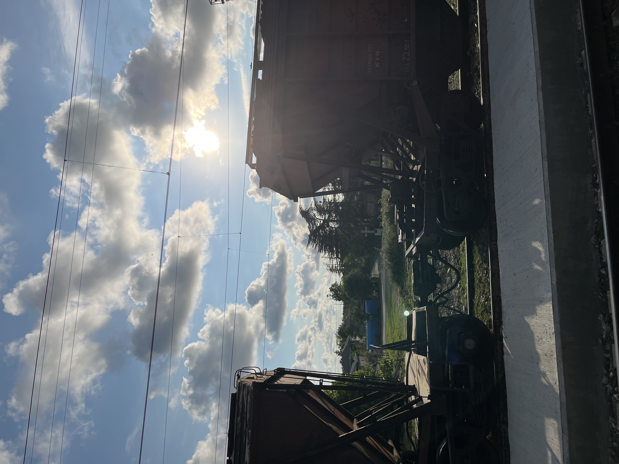 The photograph, taken in Ureki, Georgia, depicts two empty railcars parked on a set of tracks. The sky above is bright blue, dotted with fluffy white clouds, and the sun is shining through, casting a lens flare effect and highlighting the scene. The railcars are a dull reddish-brown color and appear to be of the hopper car type, commonly used for transporting bulk materials like grain or coal. One railcar is positioned on the left side of the frame, partially obscured by the other, while the second railcar occupies the right side, showing more of its details. The ground around the tracks is covered in grass and gravel, with some vegetation and scattered buildings visible in the background. A concrete platform runs along the foreground. The image captures a quiet moment at what appears to be a train station or industrial area.