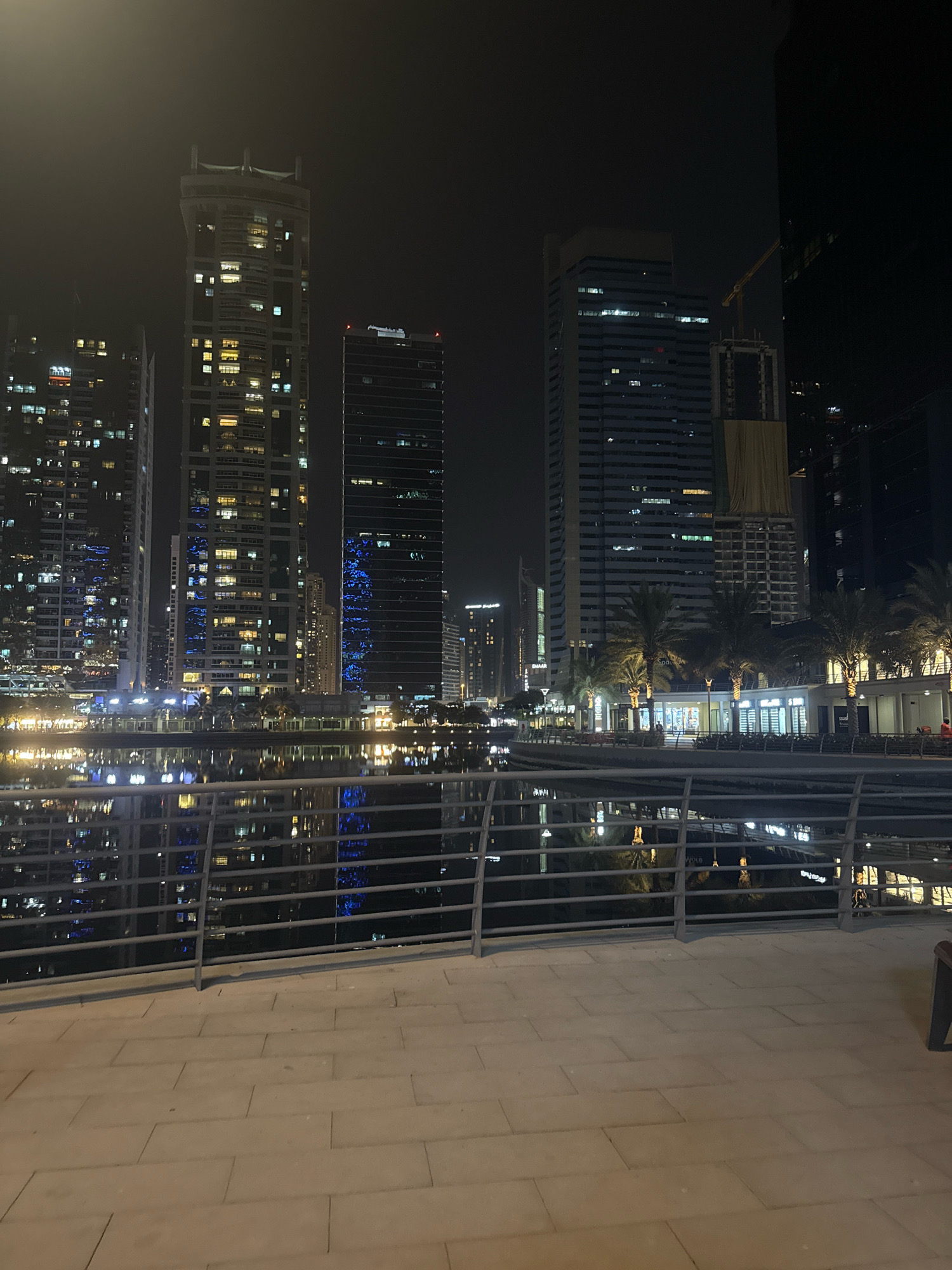 This is a nighttime photograph of a waterfront promenade in Dubai, United Arab Emirates. The foreground features a paved walkway with a metal railing, overlooking a body of water. Across the water, a line of illuminated skyscrapers dominates the background. Some of these buildings show distinct architectural features, with windows lit up in various colors, including warm yellow and bright blue. One building appears to be under construction, with a yellow crane visible above it. Palm trees line the waterfront on the far side, with lights illuminating the area. Reflections of the buildings and lights are visible in the calm water. The overall impression is of a modern, vibrant urban landscape at night.
