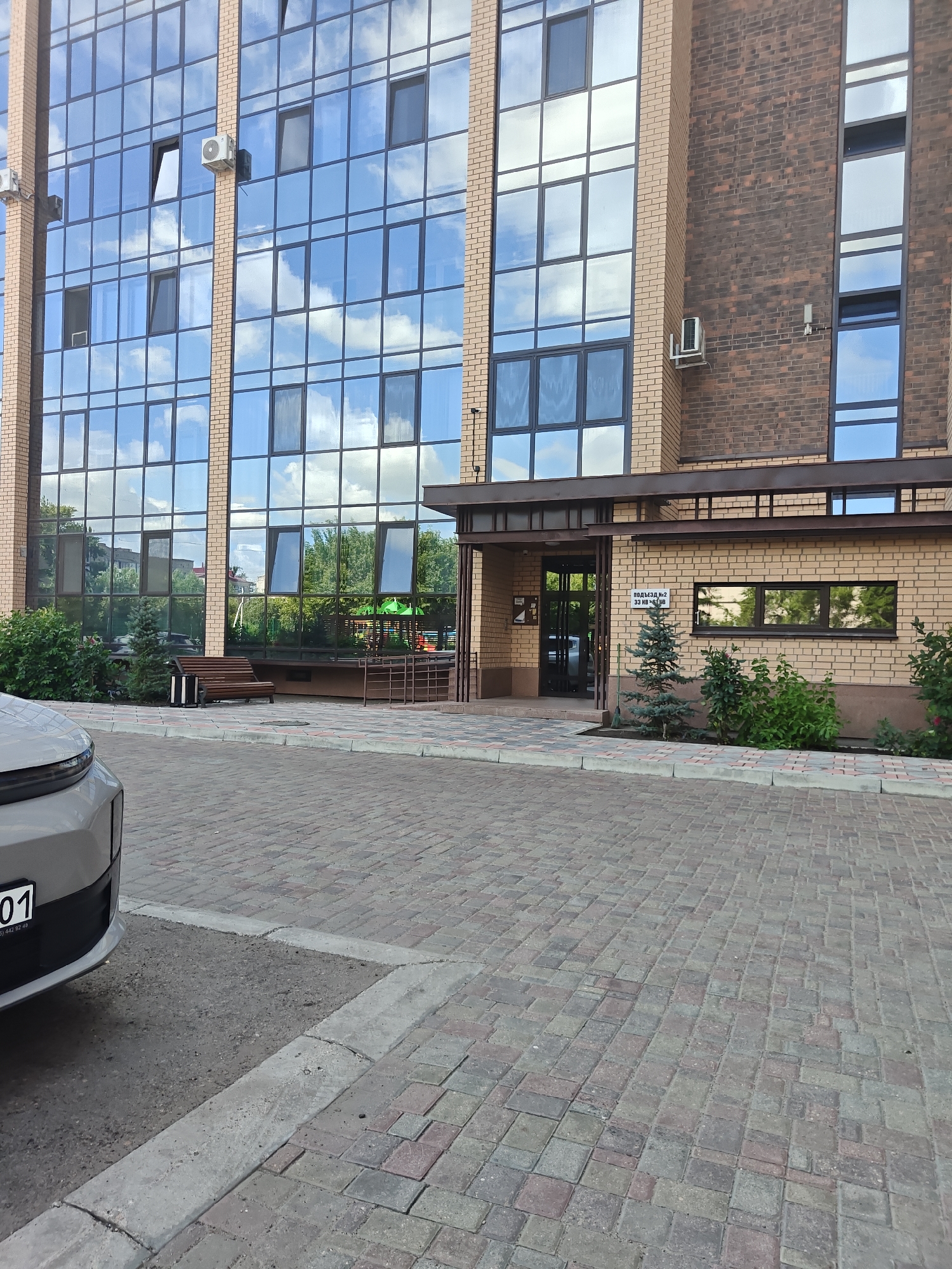 The image shows the exterior of a modern building with large glass windows and brick accents. In the foreground, a portion of a gray car is visible on a paved driveway. The building features multiple stories with reflective windows that show a clear sky with clouds, as well as greenery and other buildings reflected. There is a canopy over an entranceway with glass doors. To the left of the entrance, a wooden bench with a metal trash receptacle is placed on a paved walkway. Landscaping with bushes and small trees is present in front of the building. A sign on the brick wall near the entrance reads "ПОДЪЕЗД №2" and "53 ИНДИВИД". This is a street-level exterior view of a building.