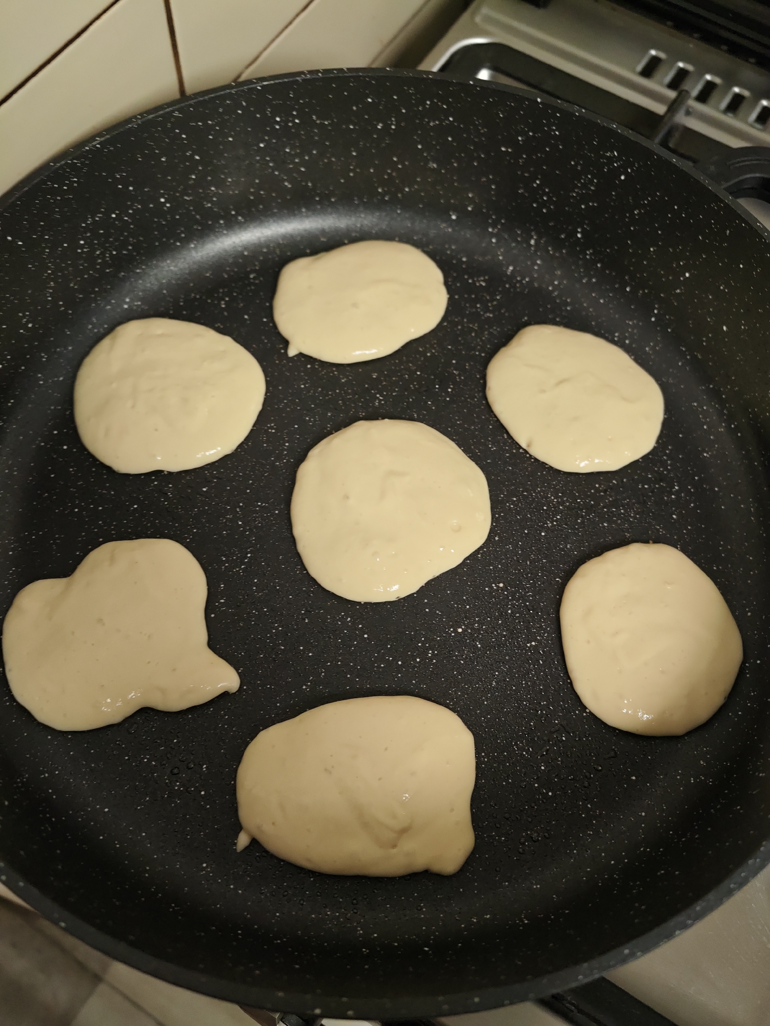 The image shows seven uncooked pancakes in a black frying pan on a stovetop. The pancakes are light golden in color and appear to be in various stages of being poured into the pan. The frying pan has a speckled pattern. In the background, a portion of a kitchen stove with silver and black elements is visible. The scene is set in a kitchen in Petah Tikva, Israel.