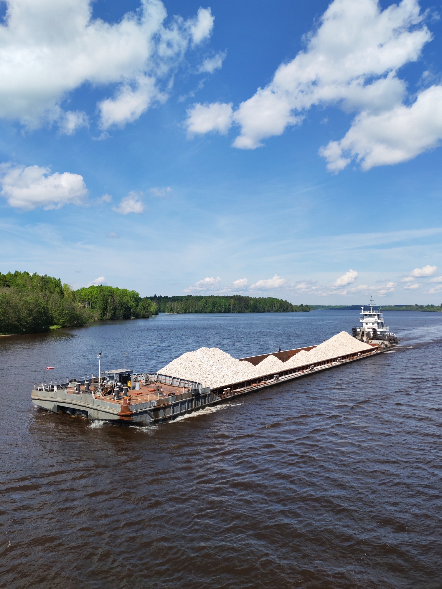 A long barge laden with a large pile of light-colored rocks is being pushed by a tugboat on a wide river. The water is dark brown. Lush green trees line the left bank of the river, while a forested area appears in the distance on the right. A blue sky with scattered white clouds is overhead. A flag is visible on the barge, and there are various pieces of equipment and structures on its deck. This scene may be in Kurikaevo, Russia, although there is no direct, verifiable information to confirm this location.