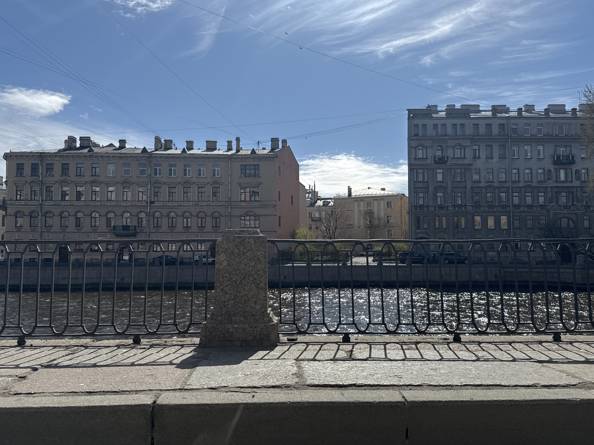 The image depicts a canal in Saint Petersburg, Russia, framed by a granite embankment. A dark metal railing runs along the embankment, its pattern of arches reflected in the water's surface. A weathered stone pillar stands prominently, interrupting the flow of the railing. Beyond the canal, buildings rise, varying in color and architectural style. The sky is a clear blue with wisps of white clouds. Power lines crisscross above the buildings. The image suggests a sunny day in a historic urban setting. Cars are parked on the street behind the buildings. The water is reflecting the sun.