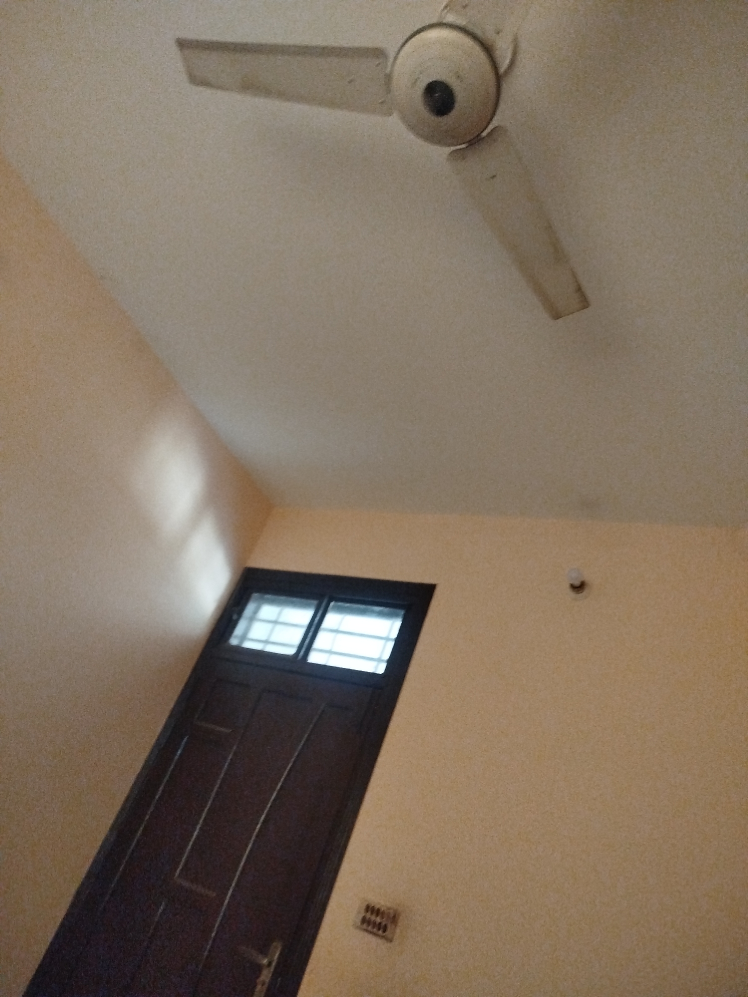 The image shows an interior shot looking up at a ceiling fan. Below the ceiling fan, a dark wooden door with a window at the top is visible. The walls are painted a light orange color. A light bulb is also visible on the wall. The scene appears to be in a room with warm lighting.