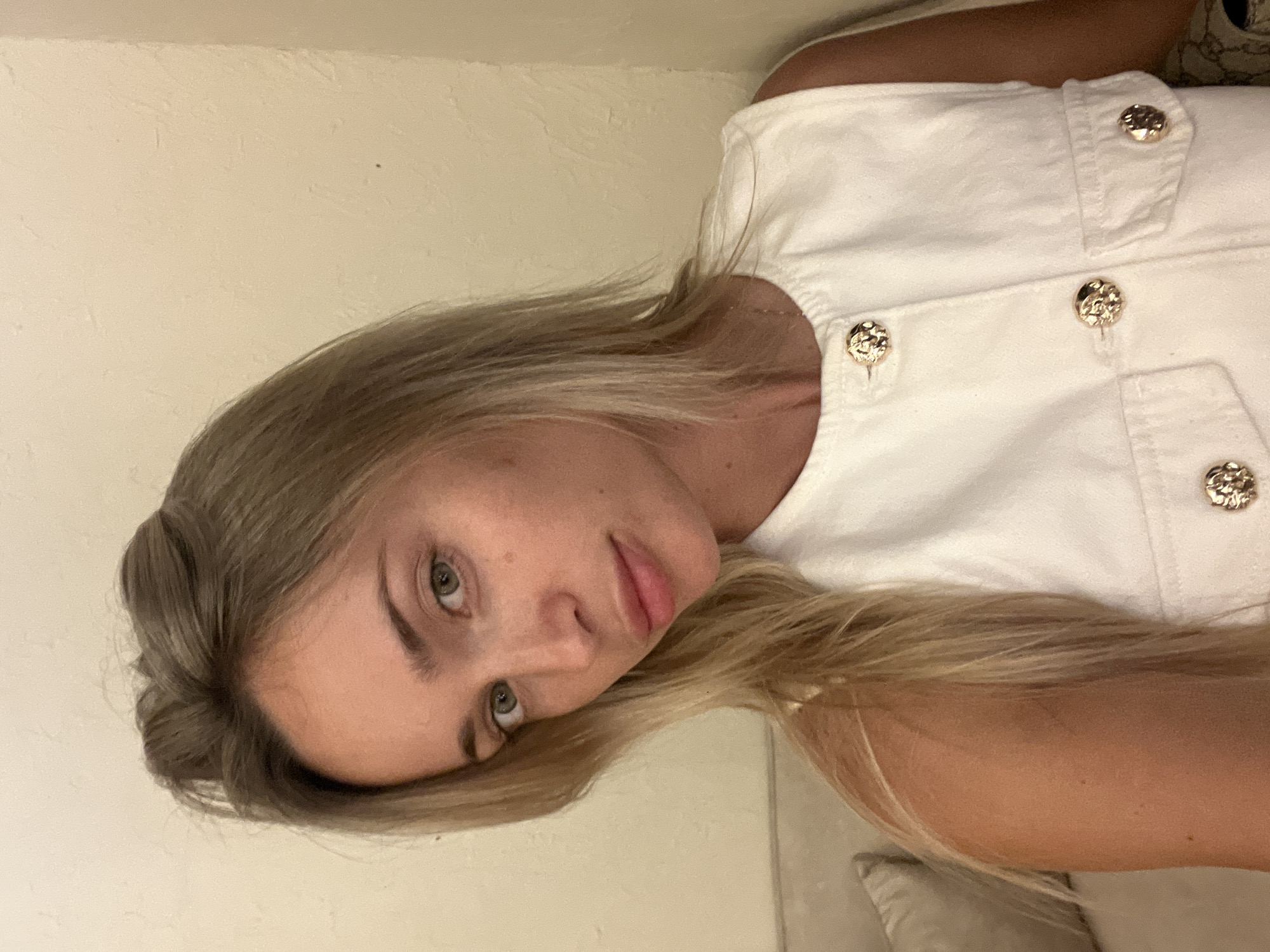 A close-up, medium shot shows a young Caucasian woman with blonde hair and green eyes. She is looking directly at the camera with a neutral expression. Her hair is pulled back from her face, with some strands falling loosely around her shoulders. She is wearing a white sleeveless top with gold-colored buttons and a subtle necklace. The background is a plain, light-colored wall, and a portion of a beige couch is visible on the left side of the image. The lighting is soft and warm, suggesting an indoor setting. No specific location in Odesa, Ukraine, can be identified from the image.