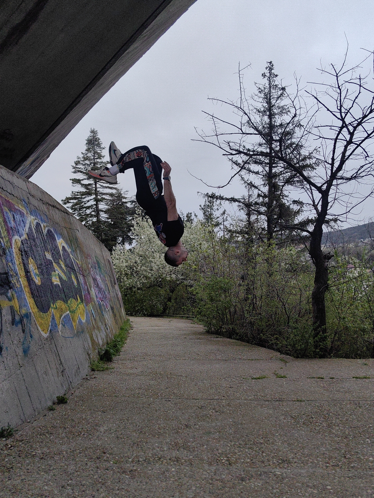 The image shows a man performing a backflip on a concrete path in Varna, Bulgaria. He is wearing a black t-shirt, patterned black pants, and grey sneakers. The path runs alongside a concrete wall covered in graffiti. Above the path is a bridge or overpass. The background includes trees, bushes, and a cloudy sky. The scene appears to be an outdoor park or recreational area.