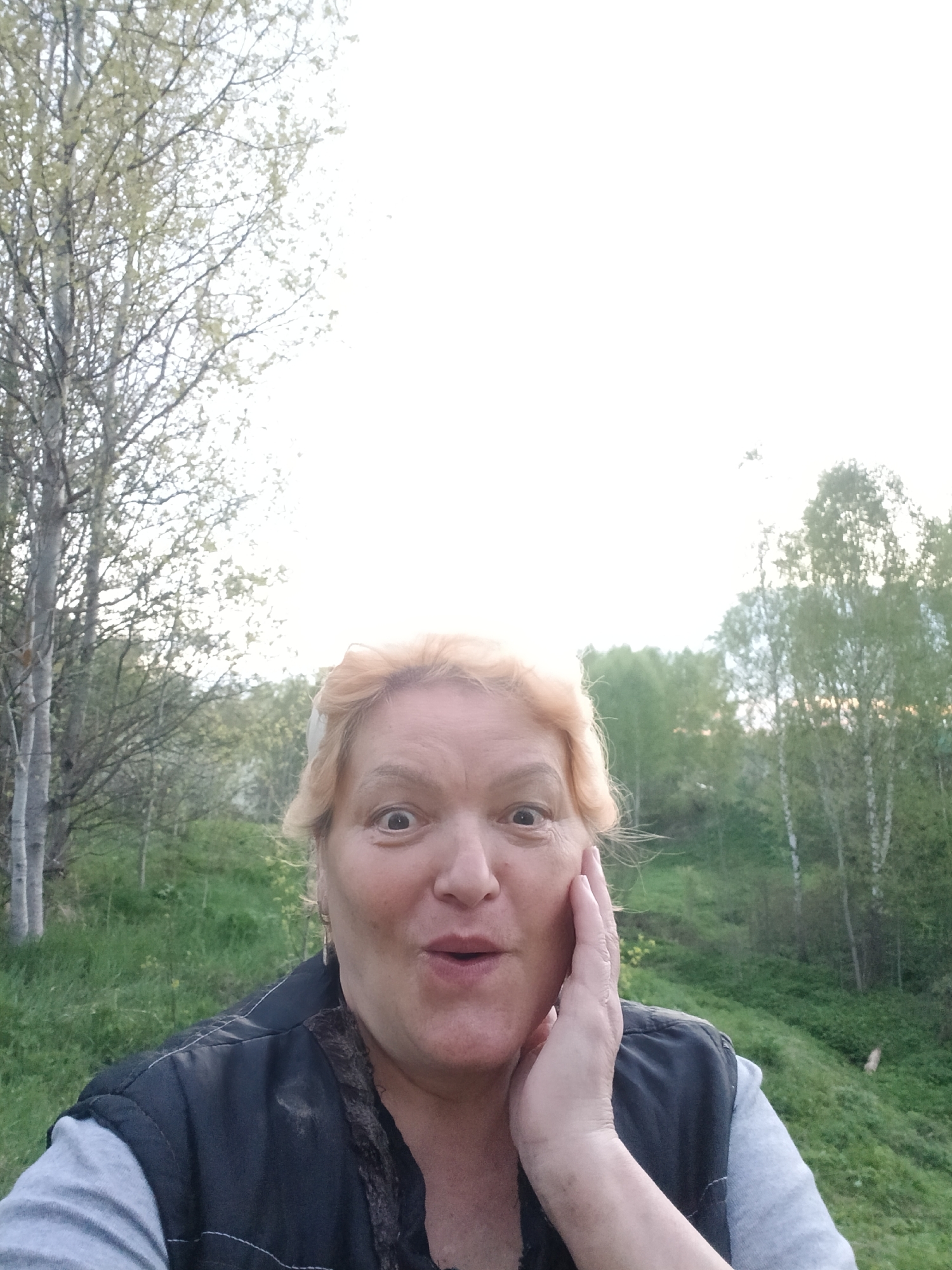 A woman stands outside in Красный Маяк, Russia. She has blonde hair and is wearing a black vest over a grey shirt. She is holding her hand to her cheek and looking at the camera with a surprised expression. Behind her are trees and green grass. The sky is bright and slightly overcast.