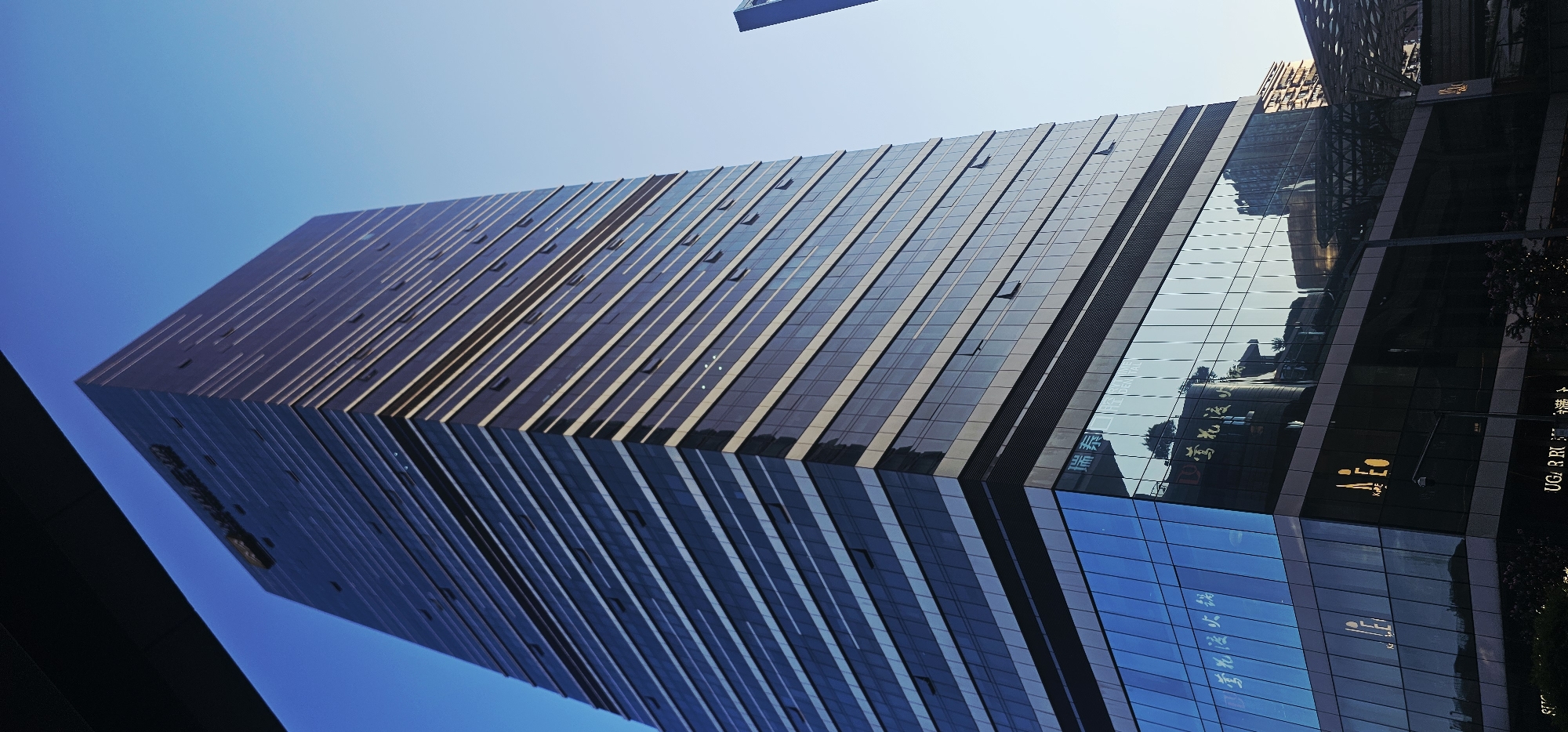 The image depicts a modern, glass-facade skyscraper in the Jiulongpo District of China, under a clear blue sky. The building is tall and angular, with horizontal bands of windows and lighter-colored trim. The lower floors of the building have a reflective glass surface that mirrors the surrounding environment, including other buildings and trees. Some signage is visible on the lower floors, including text that appears to be in Chinese characters and the word "DENTAL." The overall impression is one of urban development and commercial architecture.