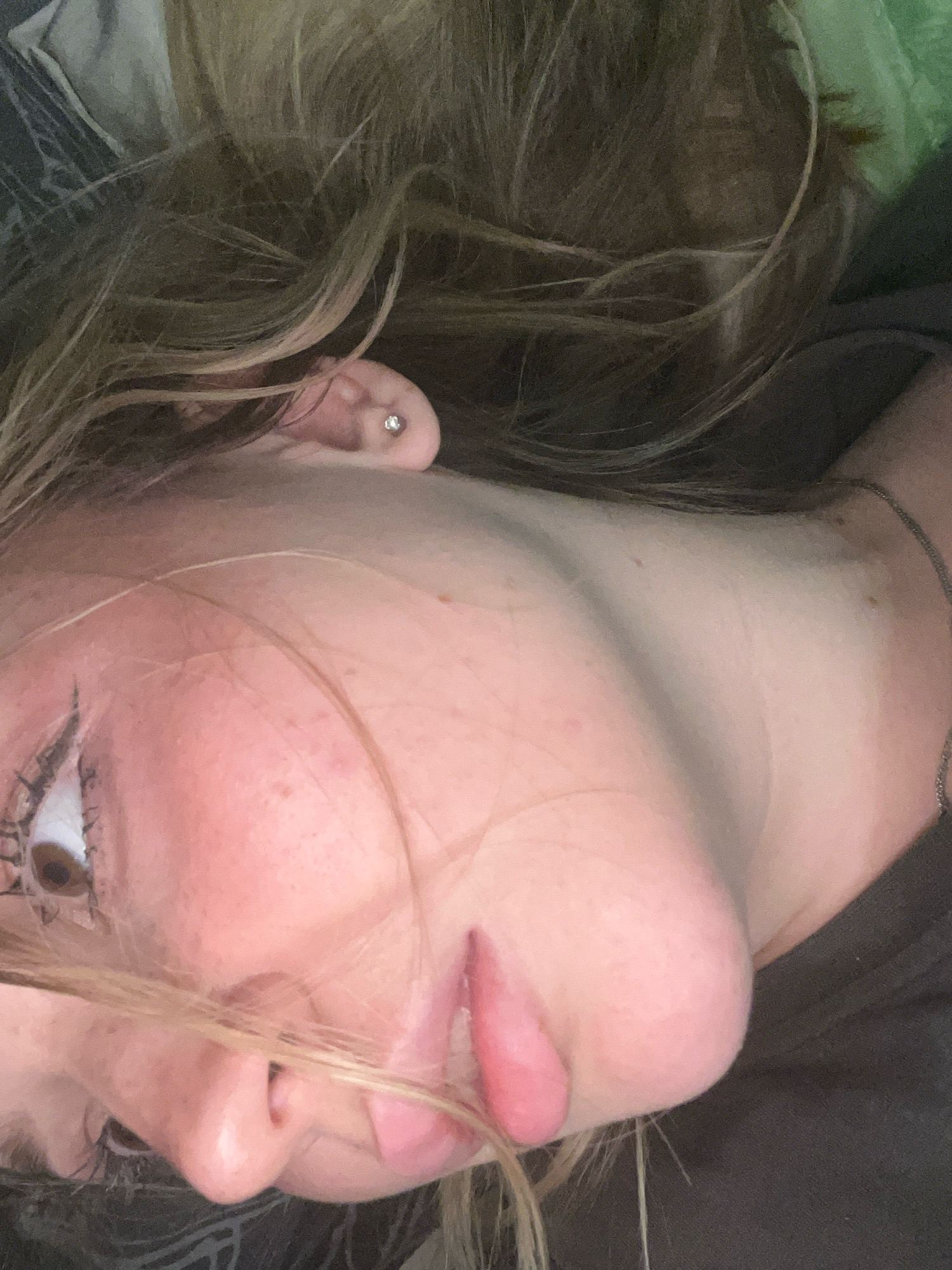 The media file is a close-up, low-angle, horizontal shot of a young person's face and upper chest, taken indoors. The person has light-colored hair that is partially obscuring their face. They are wearing a silver stud earring in their visible ear. Their skin appears fair with some redness on the cheeks and nose. The person is wearing eye makeup, including mascara. Their lips are slightly parted and appear to be pink. A silver chain necklace is visible around their neck. The background is dark and out of focus, suggesting an indoor setting with low lighting. The person's head is tilted to the side, and their gaze is directed away from the camera. The overall mood is casual and intimate. There is no visible text in the image. The location context of Nizhny Novgorod, Russia, does not provide any specific visual cues within the image itself.