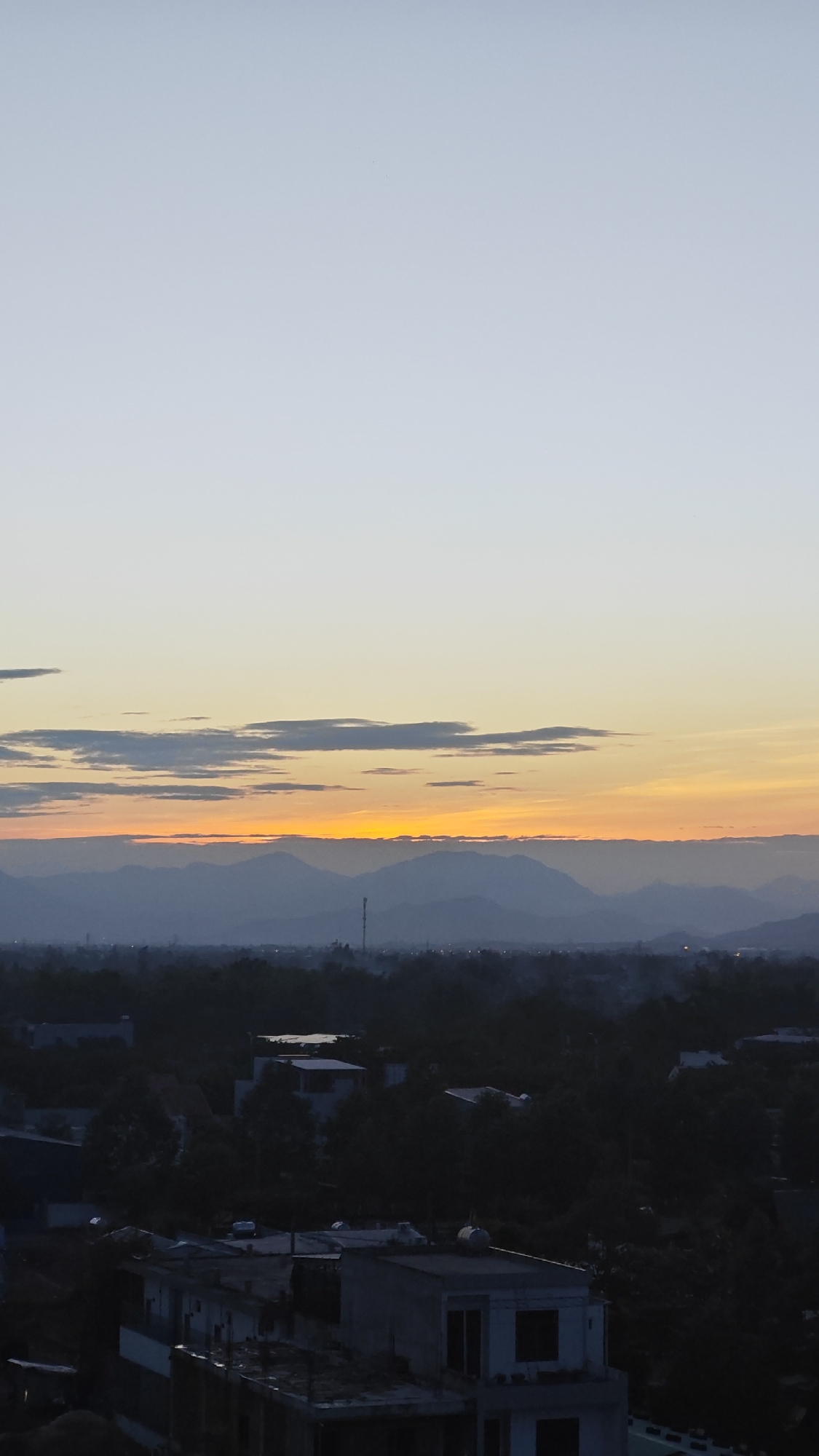 This is a vertical, eye-level, outdoor, full shot of a city skyline at dusk or dawn, taken from a high vantage point. The sky transitions from a pale blue at the top to a soft yellow and orange near the horizon, with streaks of clouds. Below the sky, a range of hazy, dark mountains forms a backdrop for the city. The city itself is composed of numerous buildings, mostly appearing as dark silhouettes against the lighter sky. Some structures are more distinct in the foreground, with visible roofs and windows, suggesting a dense urban area with a mix of residential and commercial buildings. Vegetation, in the form of trees and possibly some foliage, is interspersed throughout the cityscape. A tall, slender structure, possibly a communication tower or antenna, is visible in the middle distance. The overall atmosphere is serene and subdued, with the fading light creating a sense of depth and quietude. The location is specified as Đà Nẵng, Vietnam.