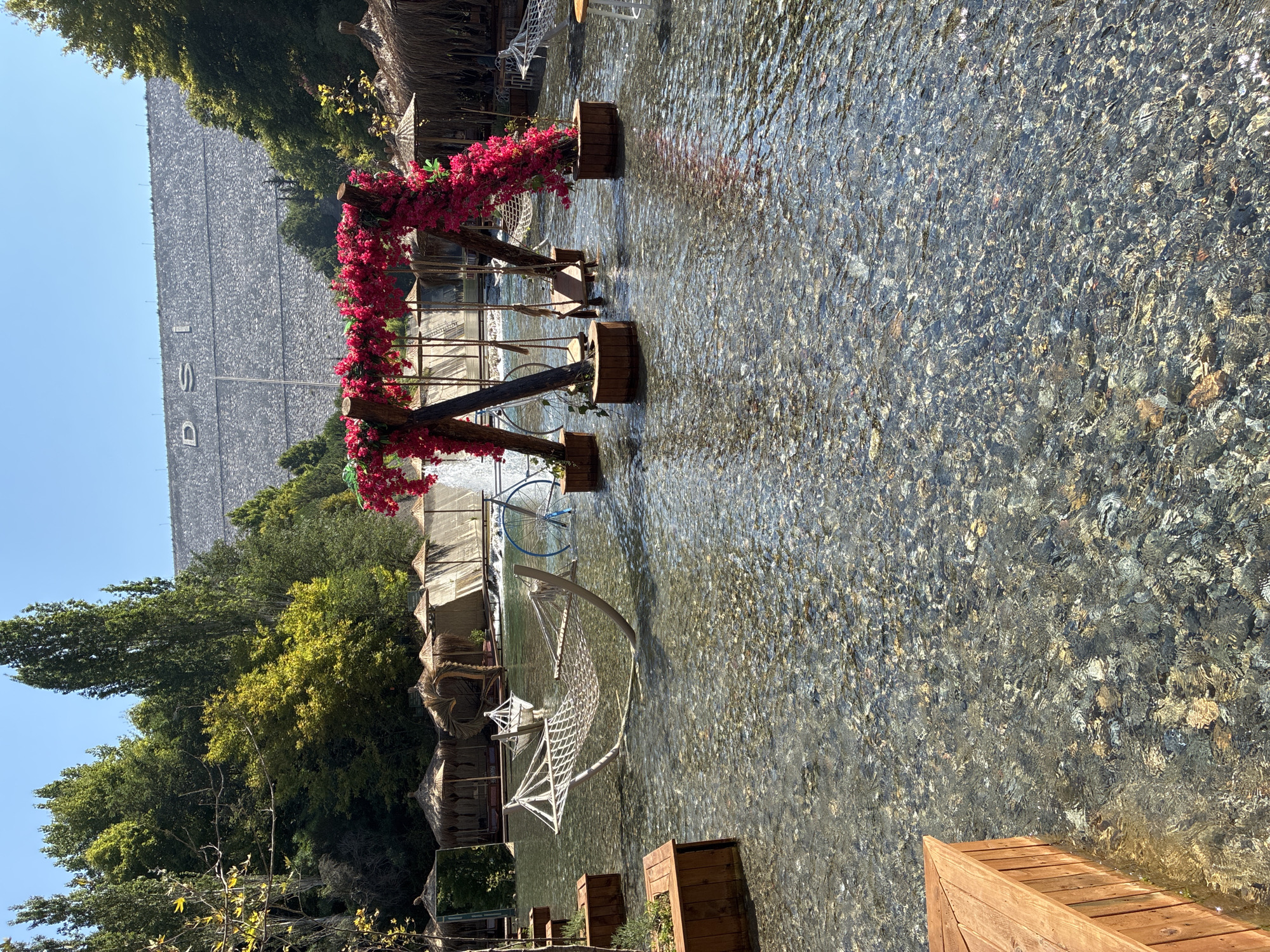 In Alanya, Turkey, a tranquil scene unfolds with a shallow, clear river serving as a unique seating area. Wooden platforms and chairs are submerged in the water, offering guests a chance to relax by the gently flowing current. Hammocks, one gracefully suspended, add to the serene ambiance.
A prominent feature of the scene is a decorative swing set, adorned with vibrant pink flowers, inviting playful moments. Behind this, a retro blue bicycle stands as a whimsical touch. Further in the background, thatched-roof gazebos provide shaded retreats.
Dominating the horizon is a massive, stone-faced dam, its imposing structure contrasting with the peaceful setting below. The dam is inscribed with the letters "DSI," a common abbreviation in Turkey for the State Hydraulic Works. Lush green trees frame the landscape, creating a picturesque backdrop. The sunlight casts dappled patterns on the water and the riverbed, enhancing the natural beauty of this idyllic spot.