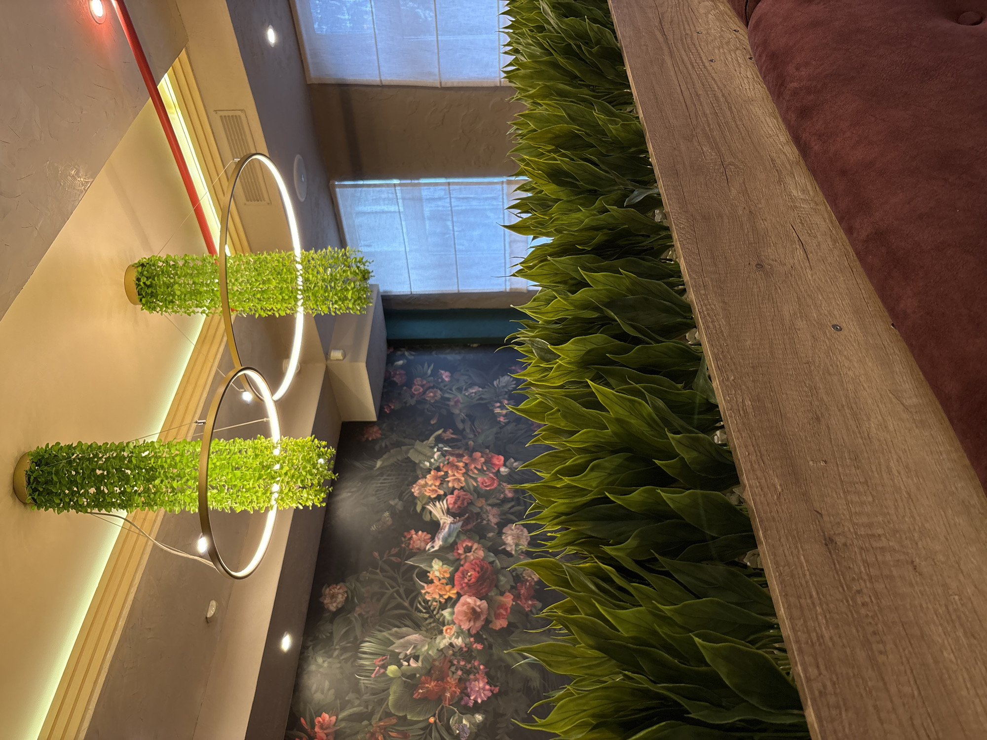 The photograph shows an interior space with various decorative elements. On the left, there are two cylindrical plant arrangements mounted on a wall and illuminated by circular light fixtures. The wall behind them features a patterned wallpaper with floral and avian motifs. In the foreground, there is a large arrangement of green plants beside a wooden structure and a maroon-colored surface, possibly part of furniture. Overhead, there are rectangular windows with white shades. Based on the prompt including the city and country name, this scene is likely an interior space within Neftekamsk, Russia.