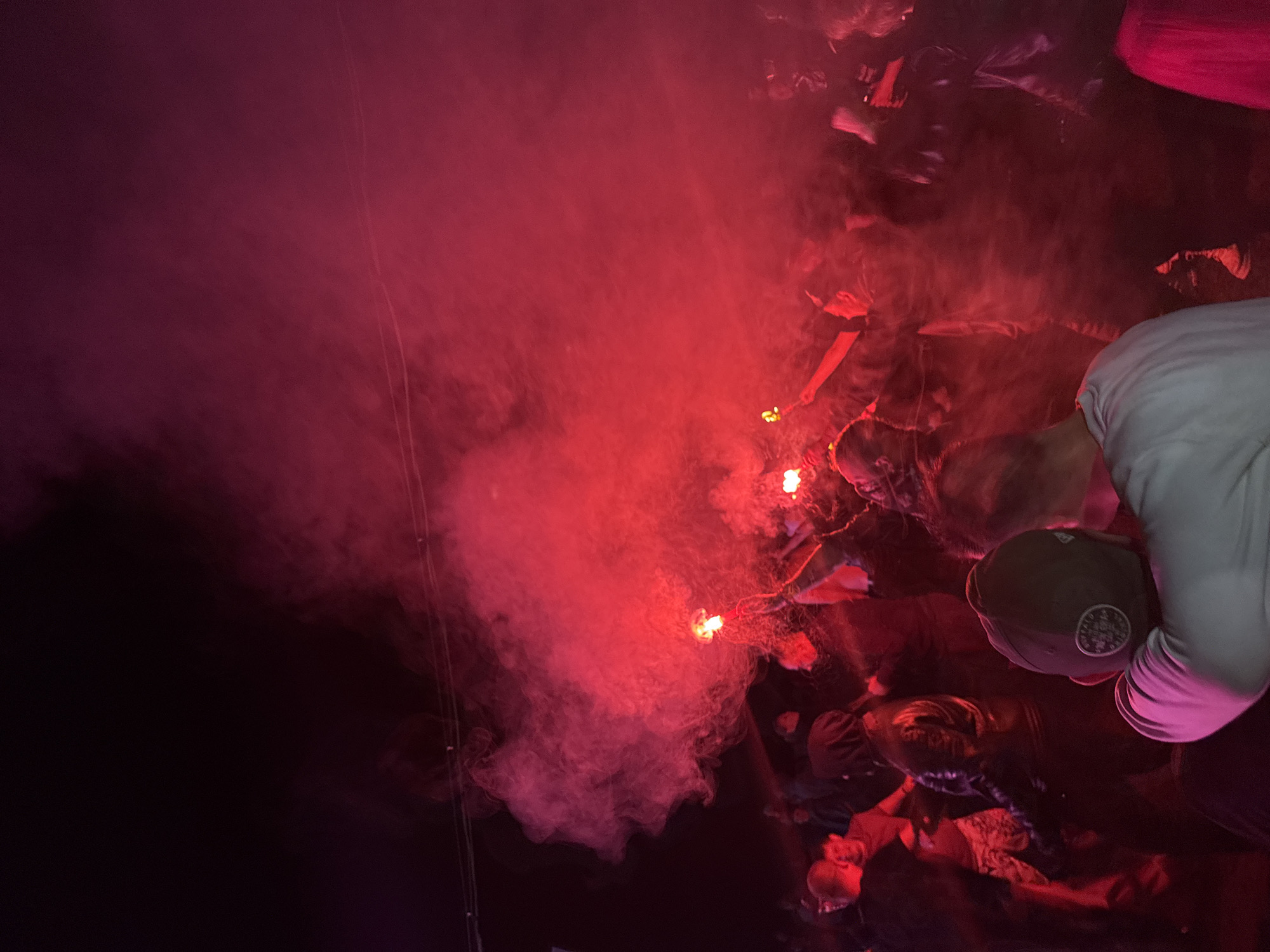 In Большая Кивара, Russia, an outdoor nighttime gathering is depicted, illuminated by strong red light and dense red smoke. The scene features numerous individuals, many partially obscured by the pervasive smoke and low light conditions. Multiple individuals are actively holding and operating red pyrotechnic flares, which are the primary source of both the red illumination and the substantial red smoke filling the upper and central portions of the frame. At least three distinct figures are observed with their arms raised, holding the burning flares. In the foreground, the backs of two individuals are visible; one wears a light-colored shirt and a dark baseball cap, positioned closely to another person. Overhead, thin wires or cables are discernible against the dark sky. The ground level is largely obscured by the crowd and smoke.
