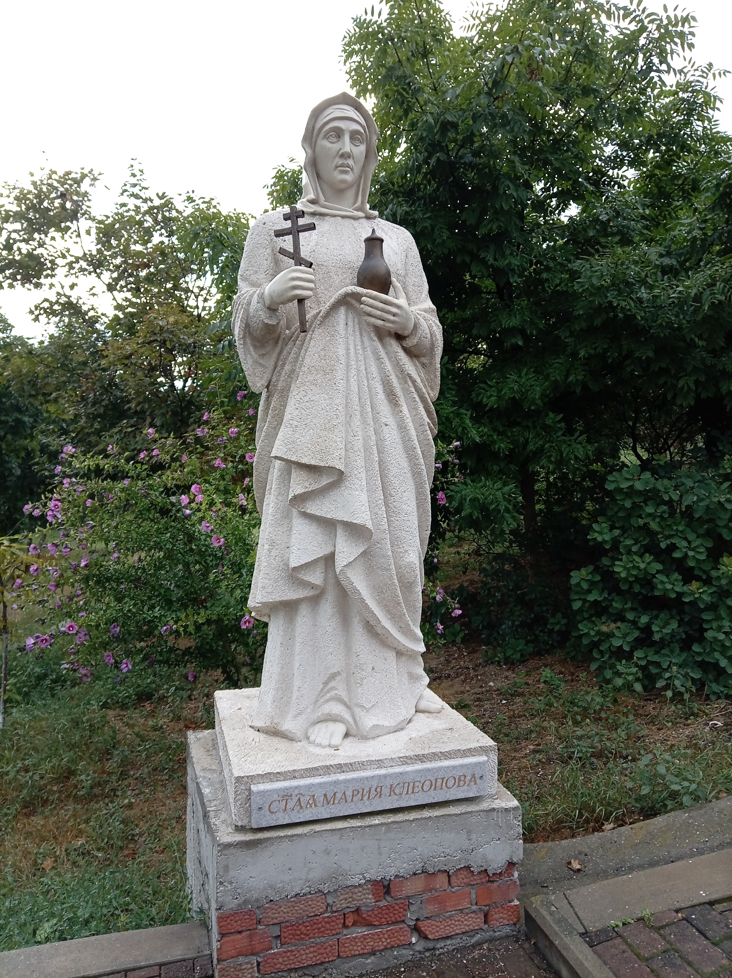 A full-body, outdoor, low-angle shot shows a white stone statue of a woman in a hooded robe, holding a cross in her left hand and a pitcher or vessel in her right. The statue is positioned on a concrete pedestal with a plaque at its base. The plaque has an inscription in what appears to be Cyrillic script. The statue is surrounded by green trees and bushes, with a few purple flowers visible in the background. The ground around the pedestal is covered in grass and dirt. The inscription on the plaque reads "СТАА МАРИЯ КЛЕОПОВА". This monument is located in the city of Yessentuki, Russia, in front of the Holy Trinity Church.