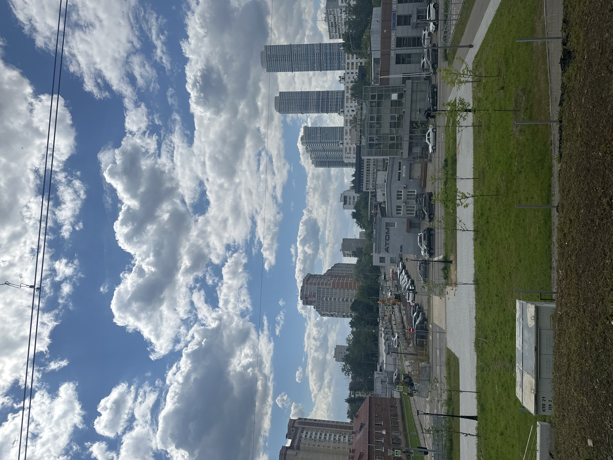 An urban street scene in Moscow, Russia, is depicted under a bright, partly cloudy daytime sky. The upper portion of the image features a clear blue sky with numerous large, white cumulus clouds, bisected by two parallel dark power lines on the left.
In the mid-ground, a cityscape unfolds with several tall, modern residential buildings featuring numerous windows, interspersed with lower commercial and residential structures. Lush green trees and bushes add natural elements to the urban landscape. Notable buildings include one with a "VOLKSWAGEN" logo and another with the word "ATOM" visible.
The foreground shows a wide, light-colored paved pathway running alongside a strip of well-maintained green grass where several young trees are planted. Beyond the grass strip, a road is visible with multiple cars, mostly light-colored, parked in rows, suggesting a car dealership or a busy parking area. A building displaying the text "АВТОСЕРВИС" and "WALKER" is also present in this area.
To the right, a dense, dark green hedge borders the scene, partially obscuring a grey electrical box. The box has visible text "КТП 21" and "ПЭ 2" on its side. The lighting suggests a sunny day with distinct shadows. No specific human activities are clearly depicted, but the environment indicates a bustling urban setting.