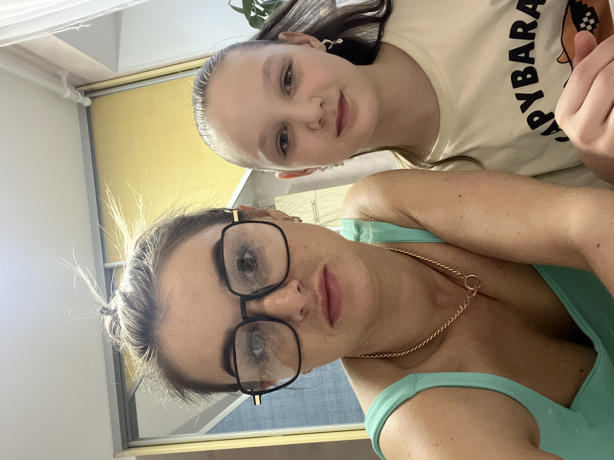 Two females are captured in a selfie-style shot, likely indoors. The adult female in the foreground wears black-framed glasses and a light green tank top with a gold chain necklace. The younger female in the background is wearing a light-colored t-shirt with black lettering.
The location is identified as Chaikovsky, Russia, although no elements in the image are definitive proof of this. A window and curtains are visible in the background. The light is bright and evenly distributed.