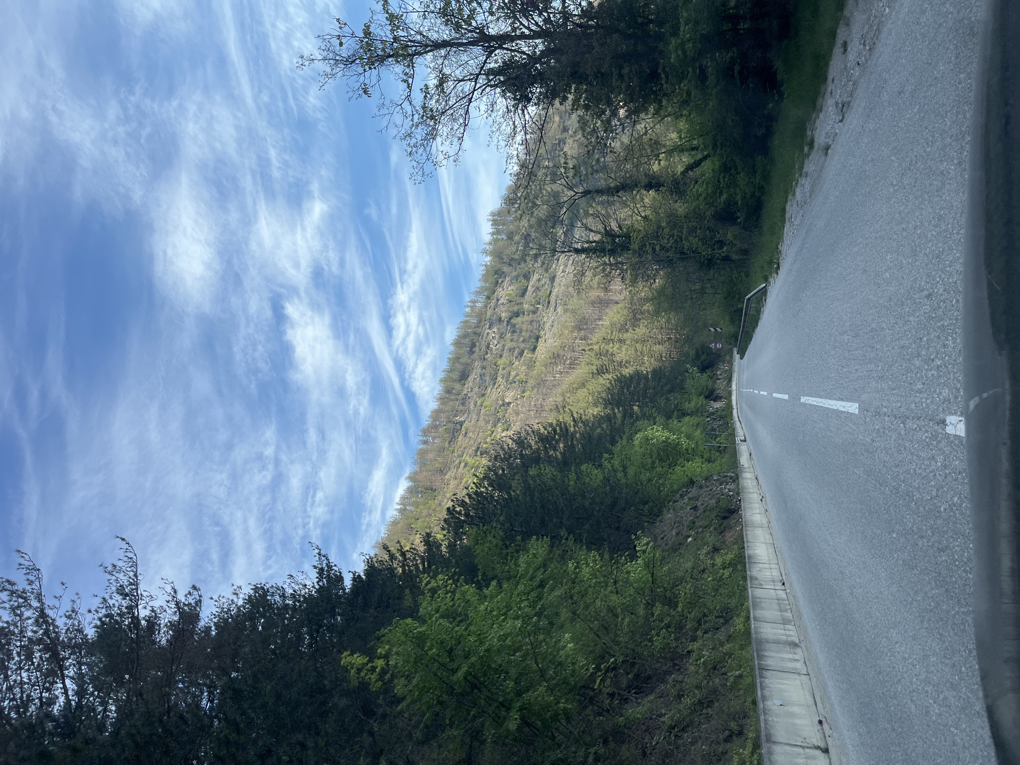 The image depicts a road in Hvoina, Bulgaria, curving alongside a forested hillside under a blue sky with cirrus clouds. The road is asphalt with white lane markings and a concrete barrier. The hillside is covered with trees and vegetation, with a mix of bare branches and green foliage.