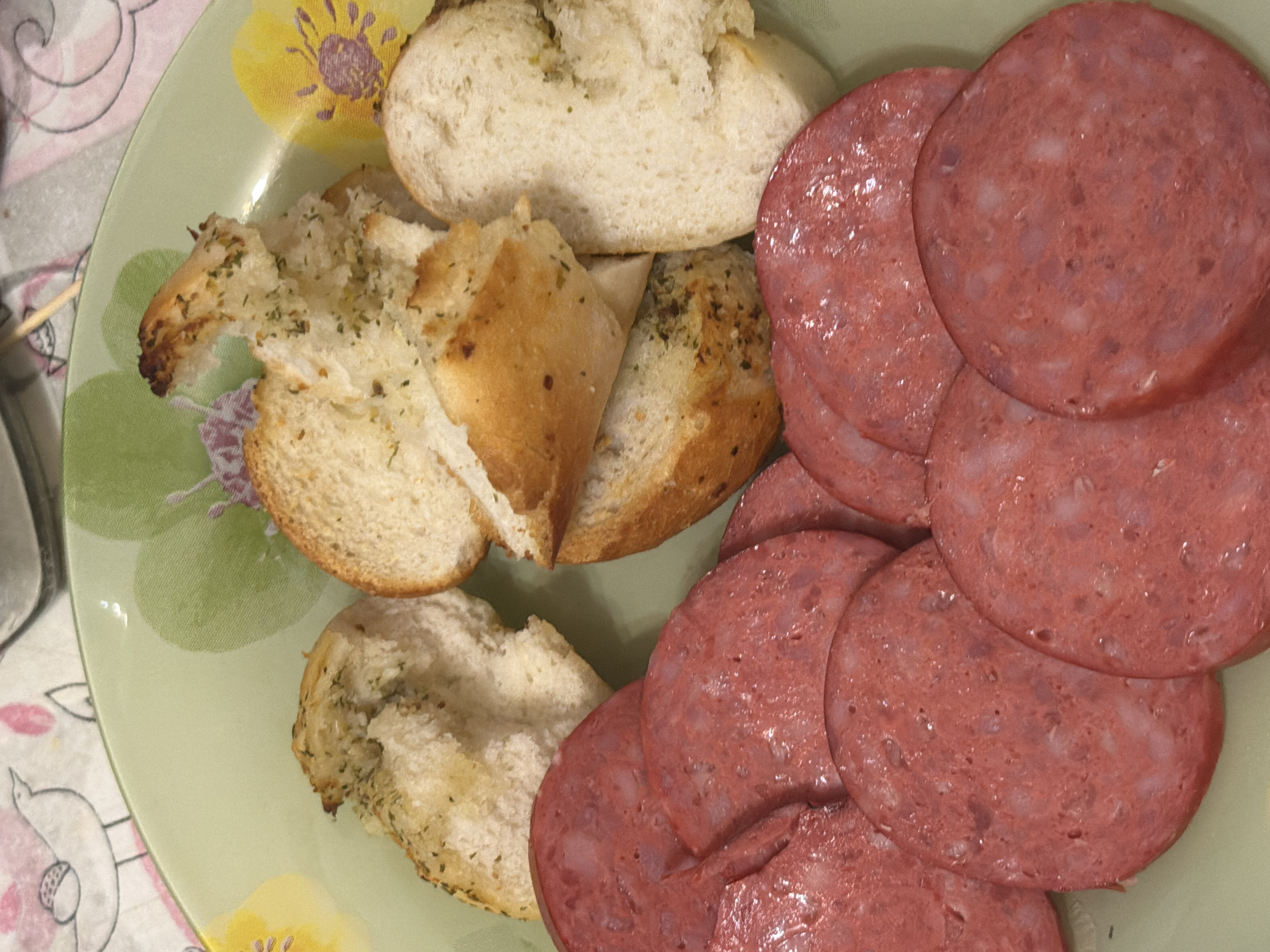 A close-up, top-down shot shows slices of salami and pieces of garlic bread arranged on a light green plate. The salami slices are round and have a reddish-pink color with visible white fat marbling. The garlic bread pieces are torn and appear to be seasoned with herbs and spices. The plate has a floral pattern with green and yellow accents. The background is out of focus, revealing a tablecloth with a bird and floral design. The lighting suggests an indoor setting.