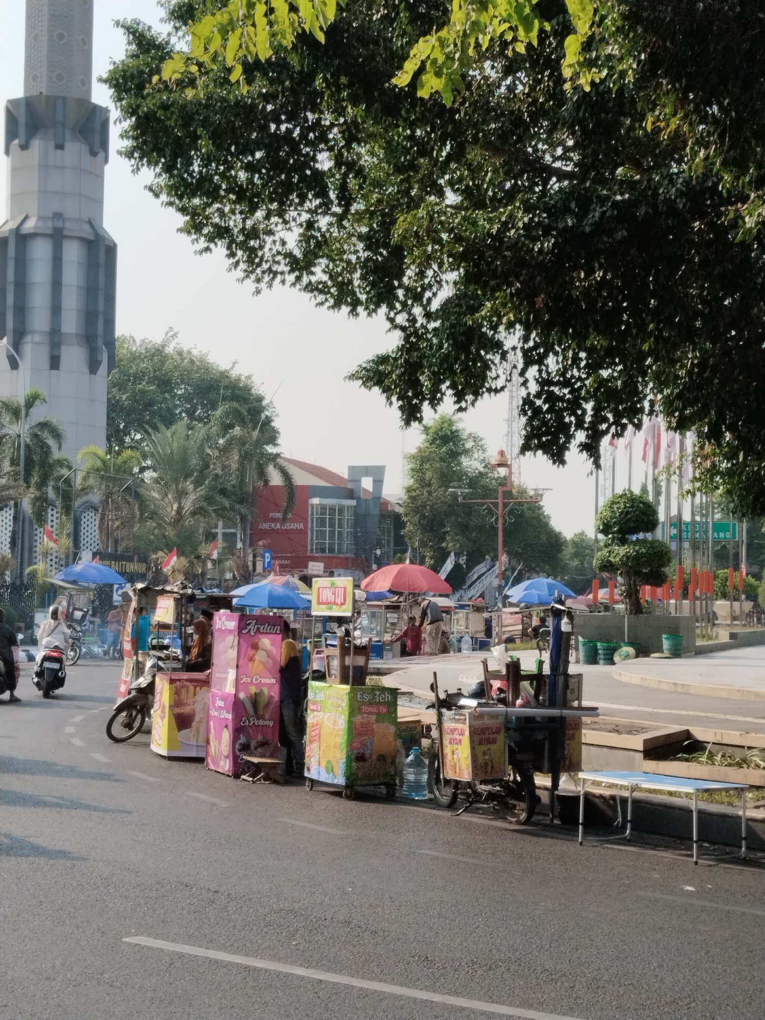 The image depicts a street scene in Kecamatan Pati, Indonesia, characterized by an outdoor market setup. In the foreground, several food carts are lined up along the side of a road. These carts are colorful and display various products, including drinks and possibly ice cream. Some of the text visible on the carts indicates offerings like "Es Teh" (iced tea) and "Ice Cream." Several people are seen interacting with the carts, either as vendors or customers. A person is on a motorcycle, and others are standing or walking around the market area. In the background, a large, modern-looking tower is visible, possibly a minaret or a landmark. Trees, including palm trees, surround the area, and there are buildings with signage, one reading "PERPUSTAKAAN KOTA ANEKA USAHA" (City Library Various Businesses), suggesting this is a public area. Flags are also visible in the distance. The overall environment suggests a busy, daytime scene in a commercial or public space.