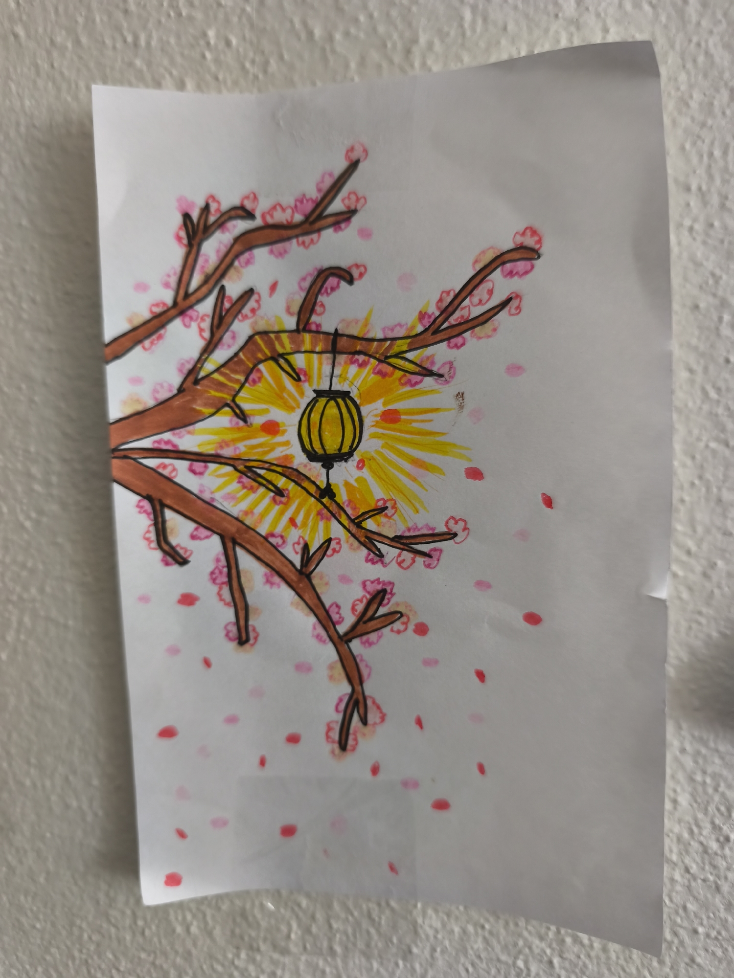 This is a photograph of a drawing on a white sheet of paper, affixed to a textured, off-white wall. The drawing depicts a stylized tree branch with pink blossoms and a yellow lantern hanging from it. The background of the drawing features radiating yellow and orange lines, suggesting sunlight or a festive light. Scattered pink petal-like shapes are also visible around the tree. The photograph is taken from a slightly elevated angle, and the right edge of the paper is slightly curled. The image does not contain any information about the city of Choceň, Czechia.