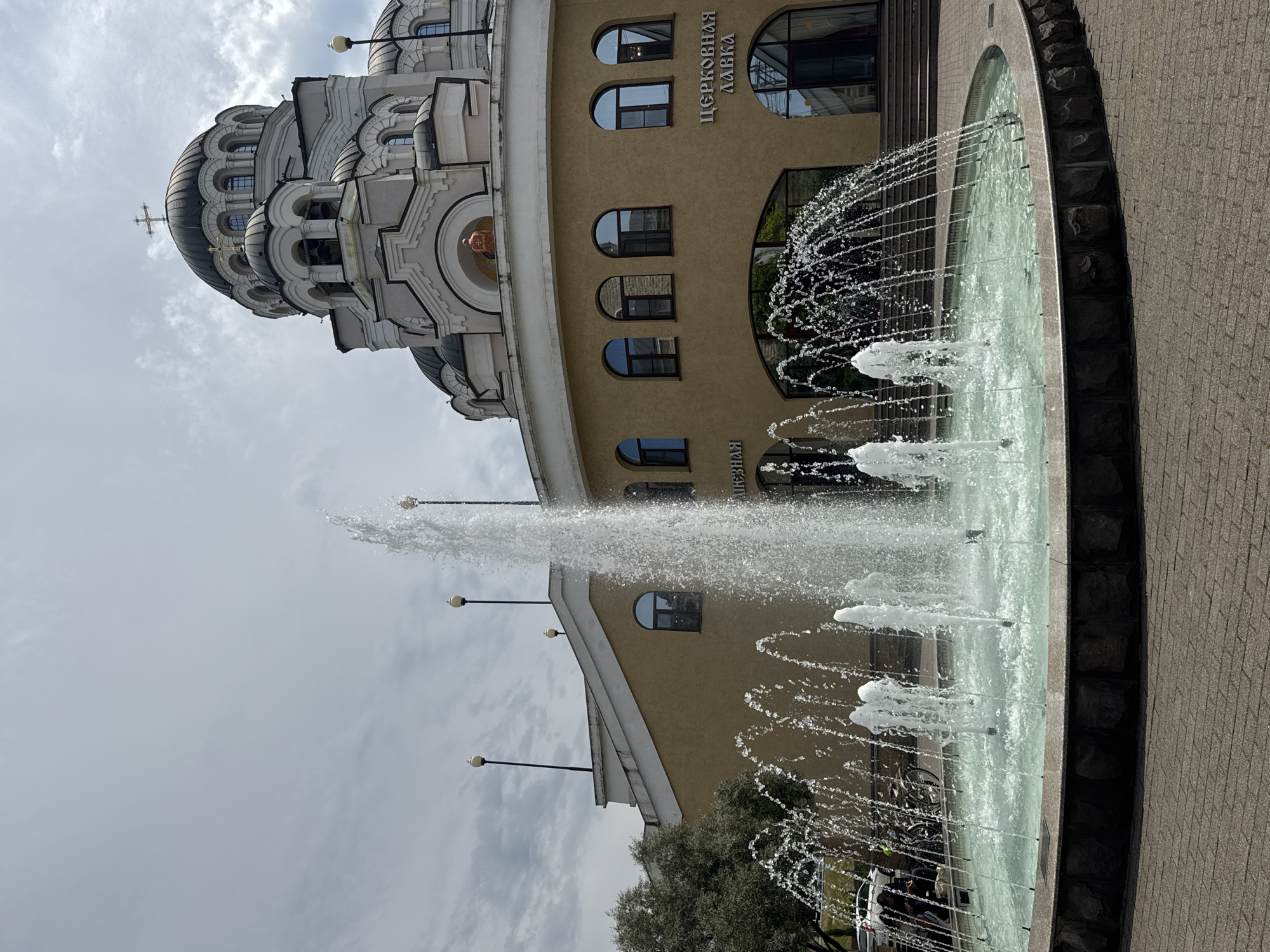 The image shows a fountain in front of a building with a church dome in Sochi, Russia. The fountain is circular, made of dark stones, and features water jets spraying upwards. The building is light brown and has arched windows. The church dome is white and gray with multiple levels and a cross on top. The sky is cloudy.