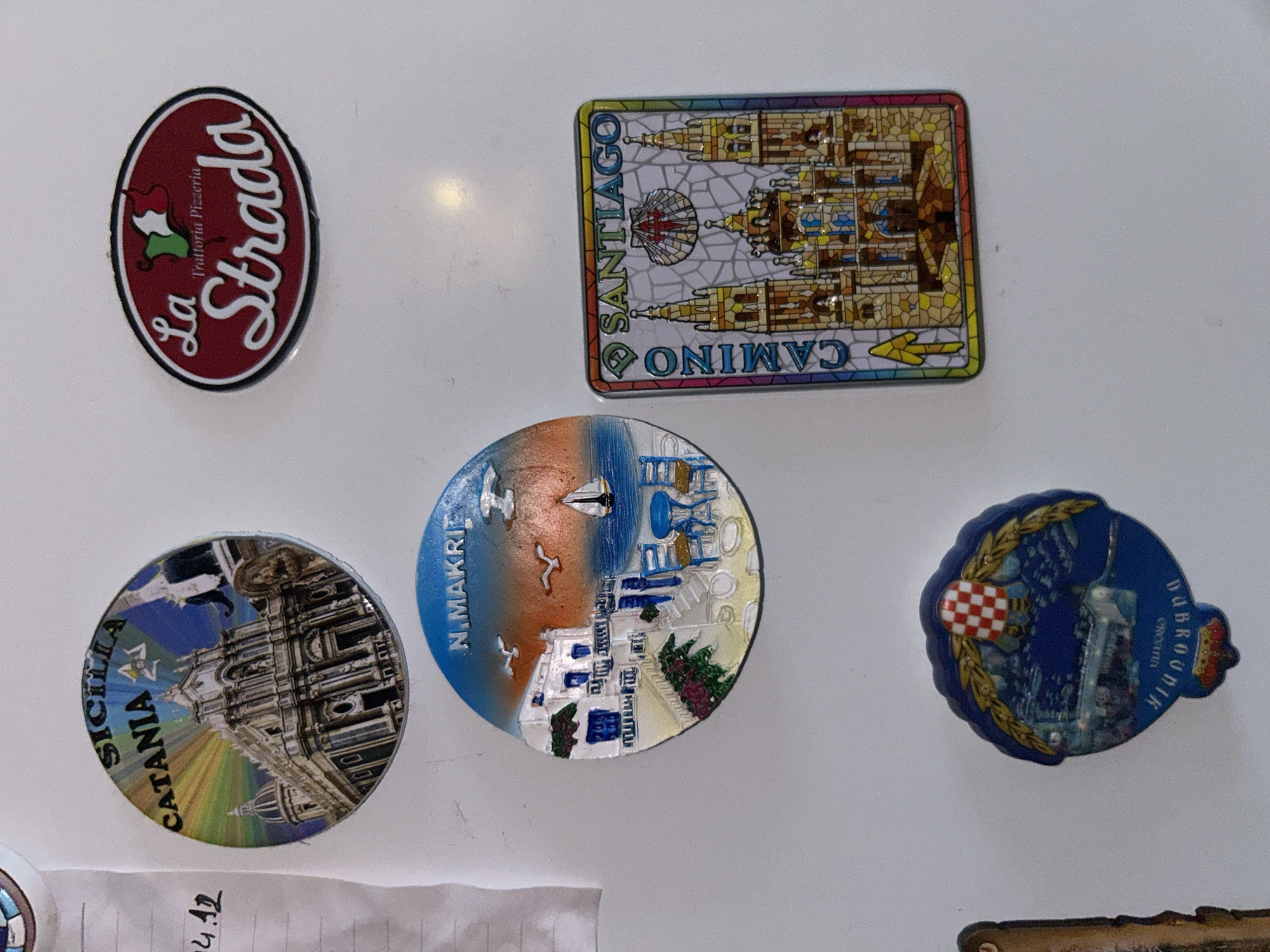 The image displays a collection of refrigerator magnets on a white surface, likely a refrigerator door. The magnets feature various landmarks and locations. From top left, a round magnet depicts a detailed architectural facade with the text "SICILIA CATANIA" and a stylized sunburst. Next to it, an oval magnet shows a red background with "La Strada" in white script, and "Trattoria Pizzeria" above it, with the Italian flag integrated into the design. Below the Catania magnet, a round magnet illustrates a white village with blue accents and seagulls against an orange and blue sky, with the text "N. MAKRI". To the right of this, a rectangular magnet features a colorful stained-glass-style depiction of a large cathedral with spires, and the text "CAMINO SANTIAGO". At the bottom left, a circular magnet with a blue border shows a nighttime scene with a cityscape and a checkered flag motif at the top, with the text "DUBROVNIK CROATIA". There are no people or discernible actions in the image. The environment appears to be indoors, with the white surface being the primary element. The specific location within the city of Sofia, Bulgaria, cannot be determined from the provided image.