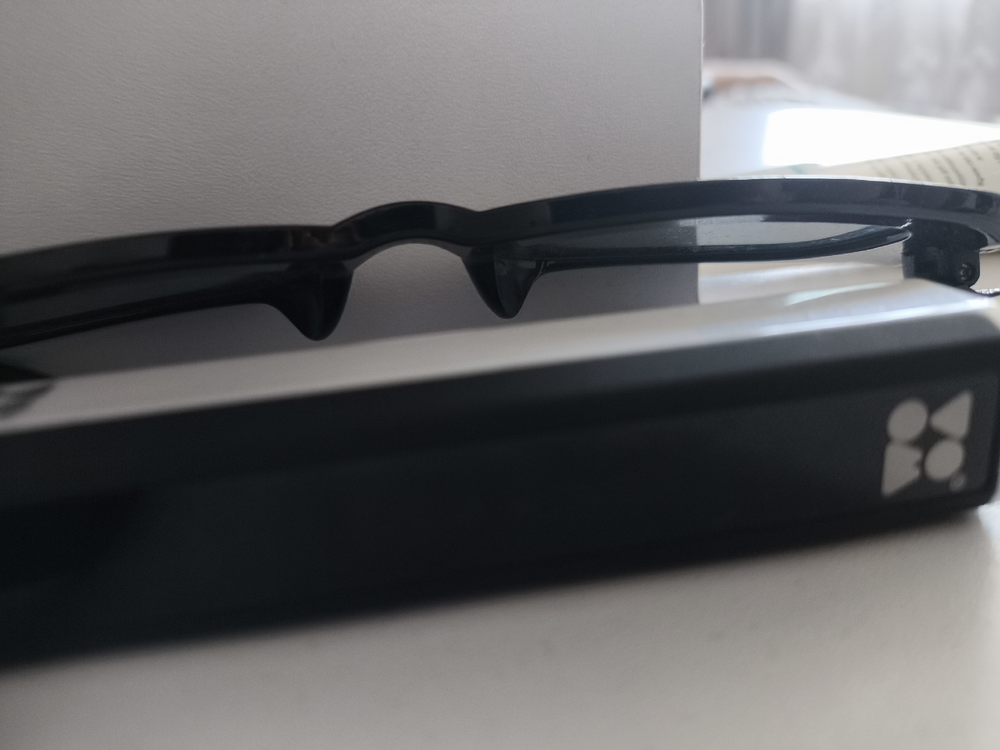 The image depicts a close-up, angled view of a pair of dark-framed glasses resting on a black, rectangular electronic device. The glasses, likely active 3D glasses or smart glasses, feature thick frames with dark, reflective lenses and integrated nose bridge components that extend downwards. They are positioned horizontally, with the front of the frames facing towards the viewer and slightly downwards, obscuring the upper surface of the device beneath them.
The black rectangular device has a smooth, matte finish. On its right end, a light grey or silver logo is visible, composed of stylized geometric shapes: two circles stacked vertically adjacent to two inverted triangles, or alternatively, a cluster of four interconnected circular or rounded elements, preceding what appears to be the letters "BO."
The scene is set indoors. The foreground is a smooth, light grey or off-white surface, likely a table or desk. The background consists of a light grey, subtly textured wall or panel. Towards the upper right of the image, a bright, diffuse light source, possibly a window, casts light onto the scene, creating a soft glow and minimal shadows. No people are visible.