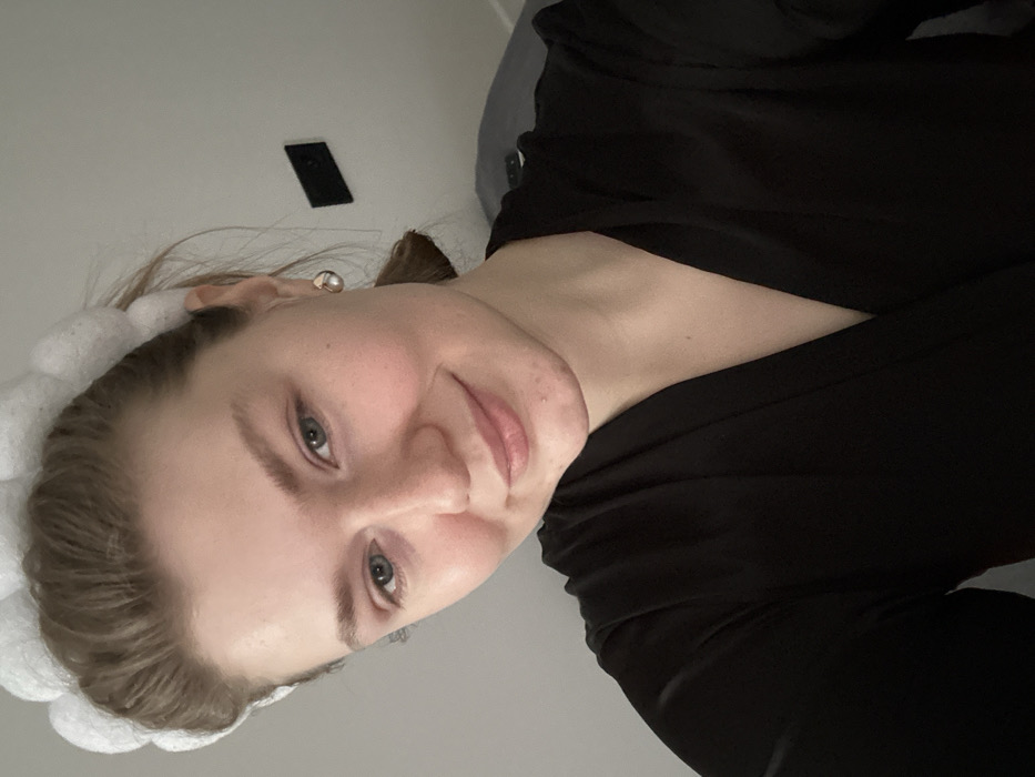 The image displays a close-up, head-and-shoulders portrait of a female individual in an indoor environment. The subject has light skin, light-colored eyes, and light brown hair pulled back, secured by a white, textured headband featuring a bubble-like design. She wears a black long-sleeved top or robe with a V-neckline and small, silver-colored stud earrings. The individual is looking directly at the camera with a subtle smile. The background consists of a plain, light-colored wall. A dark, rectangular object, resembling an electrical outlet or light switch, is mounted on the wall behind her right shoulder. An indistinct dark object, possibly bedding or furniture, is partially visible to the right of the frame. The specific location within the city of Kazan, Russia, is not identifiable from the visual information presented.