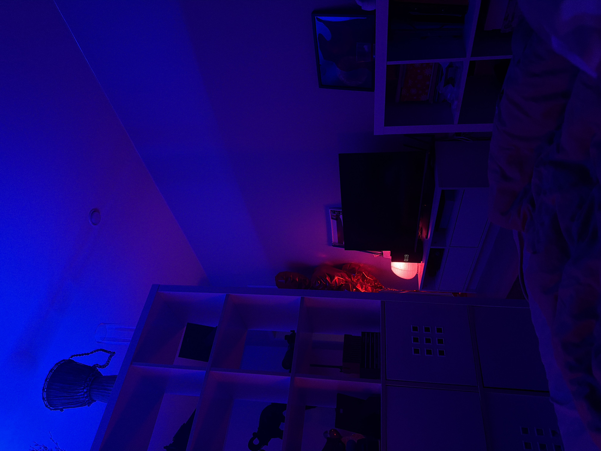 The image depicts the interior of a room, likely a bedroom, under intense blue and purple lighting. The perspective is tilted, with the ceiling appearing on the left side. The room contains shelving units, a television, a red light fixture, and assorted decorative objects, including what appears to be a djembe drum. There are no people visible in the image. Given the objects and lighting style, it appears to be a personal space. There is no information to indicate this photo was taken in Krasnoyarsk, Russia.