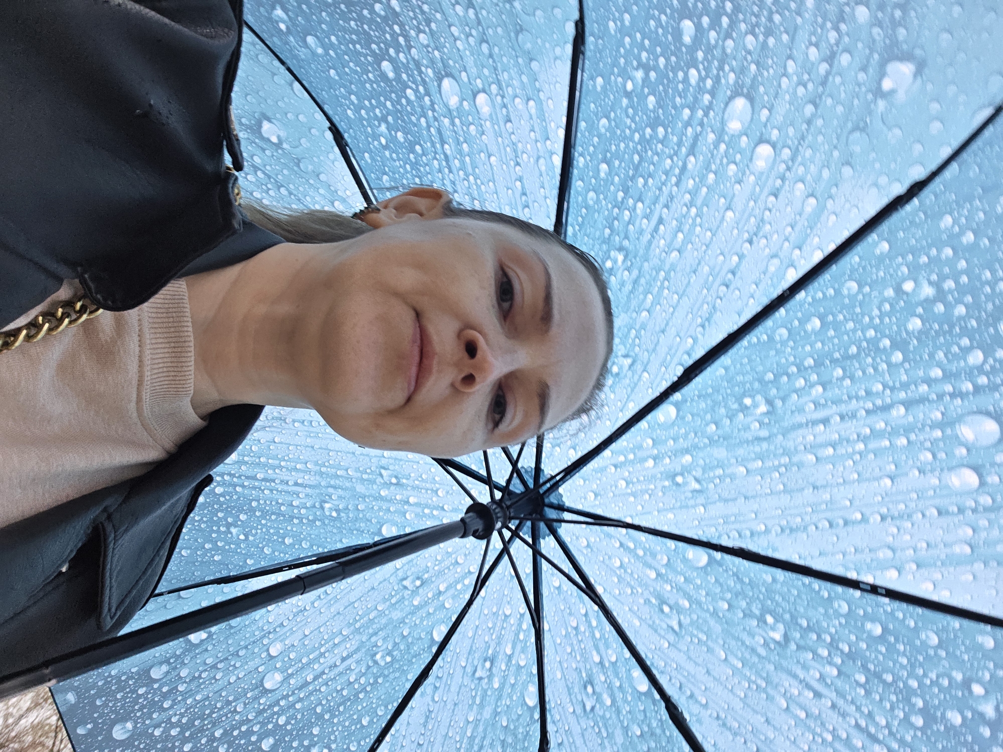 The image captures a unique low-angle perspective looking up at a woman holding an open umbrella during rainfall. The woman's face is positioned in the upper center-left, with her looking downwards towards the camera. She has light-colored hair pulled back, and a neutral to slightly pleasant expression with a hint of a smile. She is wearing a light-colored, possibly beige or light pink, ribbed top and a dark (likely black) jacket, with a visible gold chain strap on her left shoulder.
Dominating the rest of the frame is the underside of a light blue or cyan umbrella, covered extensively with numerous clear water droplets of varying sizes. This detail clearly indicates that it is raining. The black spokes and central mechanism of the umbrella are visible, radiating outwards from a point near the bottom center of the image. The overall setting is outside, in Chaikovsky, Russia, during what appears to be daytime, experiencing wet weather. There is no visible text in the image.
