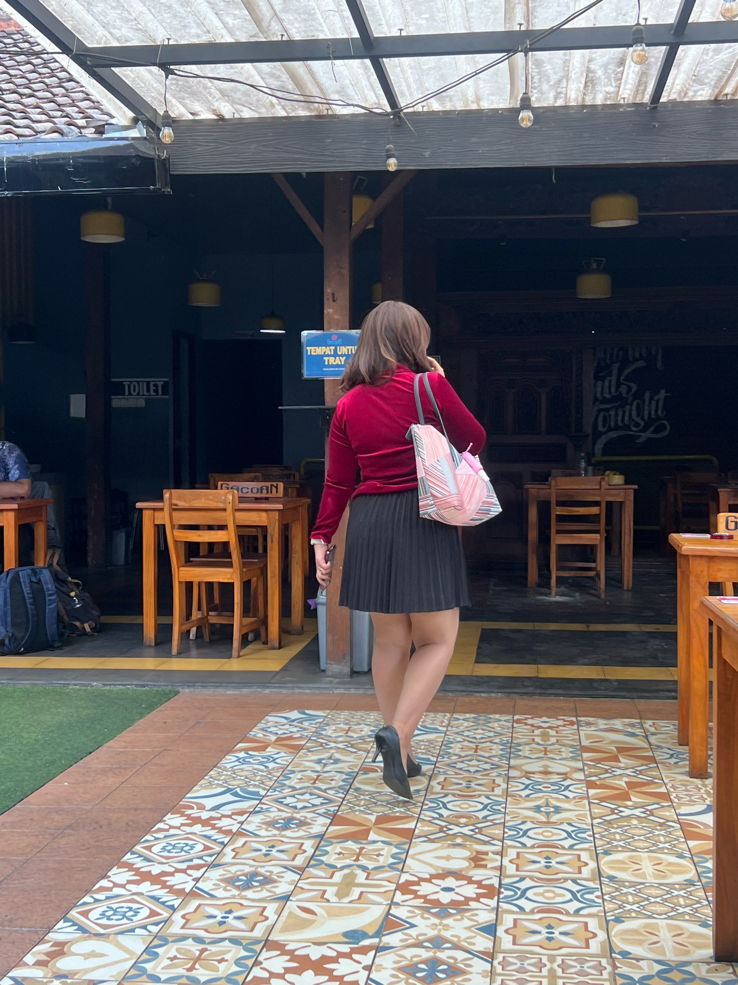 A woman with medium-length brown hair, seen from behind, walks away from the camera on a patterned tiled floor. She is wearing a red velvet long-sleeved top, a black pleated skirt, and black high-heeled shoes. A colorful tote bag with pink, white, and blue stripes is slung over her left shoulder. She is walking towards an outdoor seating area of a building with a covered roof supported by wooden beams. In the background, there are several wooden tables and chairs. A sign that says "TEMPAT UNTUK TRAY" (Place for Tray) is visible. To the left of the image, a sign reads "TOILET." The environment suggests a cafe or restaurant in Yogyakarta, Indonesia, possibly with a rustic or traditional design, as indicated by the wooden elements and the patterned tiles. The floor transitions from the patterned tiles to a reddish-brown tiled walkway and then to a patch of artificial grass.