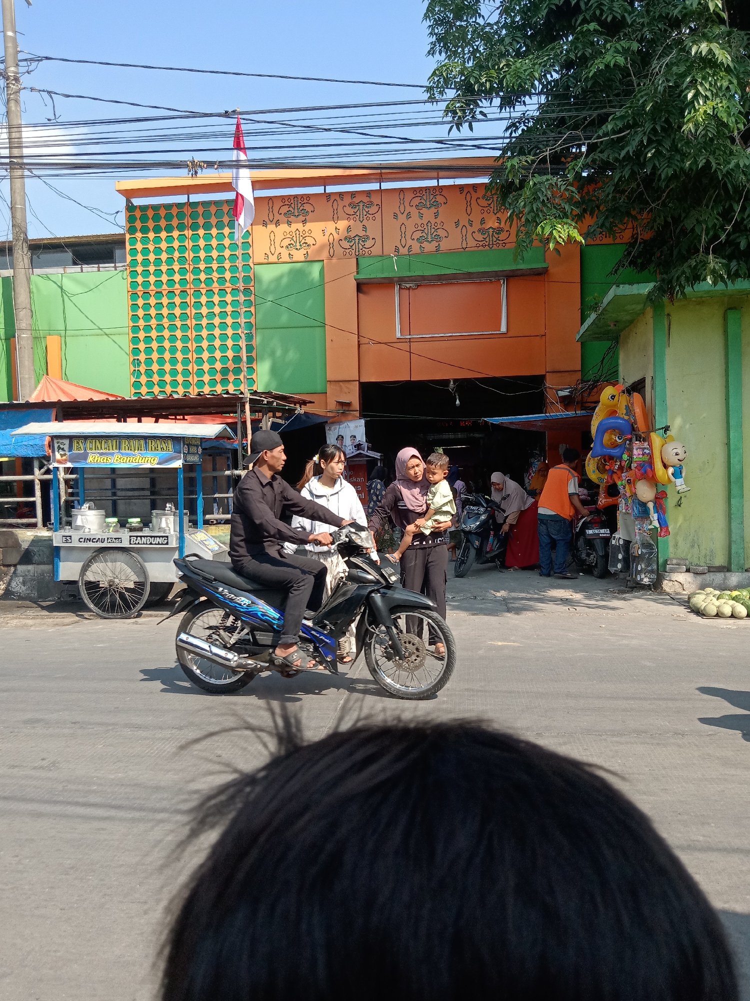The image captures a street scene in Sugihwaras, Indonesia, on a clear, sunny day. In the foreground, the back of a person's dark hair is visible, obscuring the viewer's perspective. Dominating the mid-ground is a man in a black shirt and pants, riding a blue and black motorcycle. He is wearing a black cap and sandals. A woman in a white jacket and light-colored pants is riding pillion behind him.
To the left of the motorcycle is a food cart labeled "ES CINC AU RAJA RASA KHAS BANDUNG," with an orange and blue awning. Several metal containers are visible on the cart. Behind the cart, a portion of a building with green and orange walls is visible, adorned with decorative patterns and a red and white Indonesian flag.
To the right of the motorcycle, a light green building features a display of colorful inflatable toys and dolls. Several people are gathered near this building, including a woman in a pink hijab holding a young child, another woman in a red skirt, and a person wearing an orange vest and blue jeans. A parked motorcycle is visible behind them. In the far right of the frame, a pile of green melons sits on the ground. The street itself is asphalt, with visible road markings. Overhead, a tangle of power lines stretches across the sky.