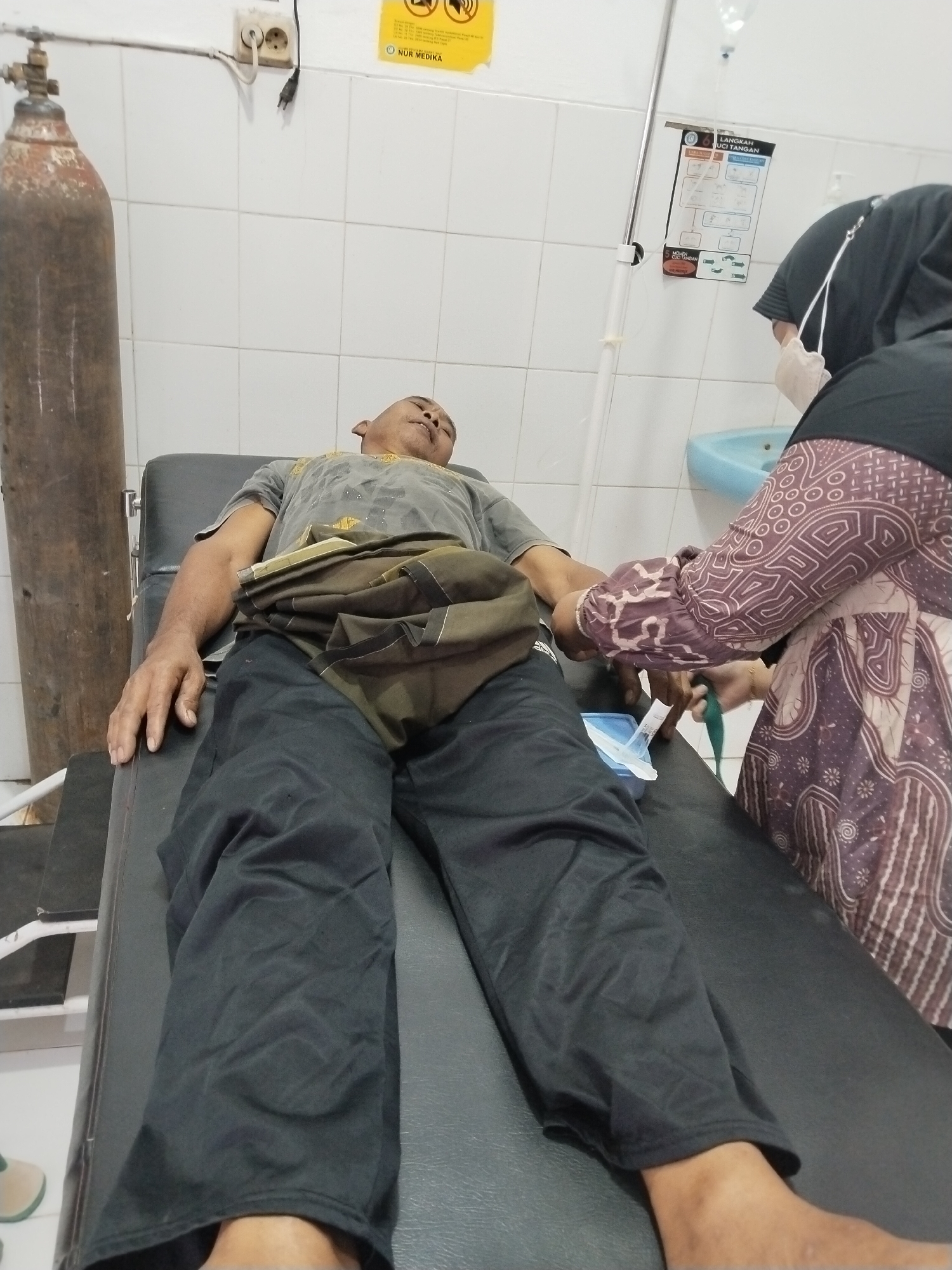 A man lies on a medical examination table with his eyes closed and mouth slightly open. He is wearing a gray t-shirt and black pants. A piece of cloth is draped over his lap. A nurse, wearing a hijab and gloves, is attending to him, with an IV bag hanging from a stand. In the background, an oxygen tank and a sink are visible. The room appears to be a medical clinic in Prigi, Indonesia, with tiled walls and a fluorescent light fixture.